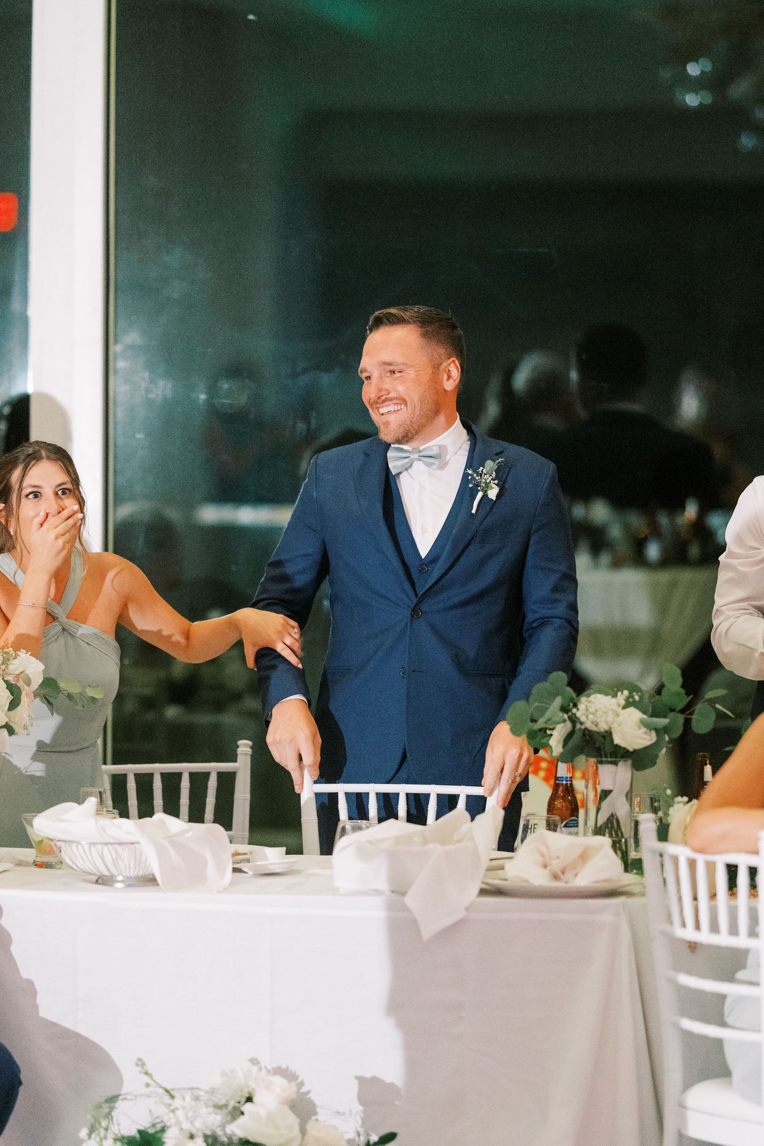 Wedding reception guests laugh and share emotional reactions at tables decorated with white linens and floral centerpieces.