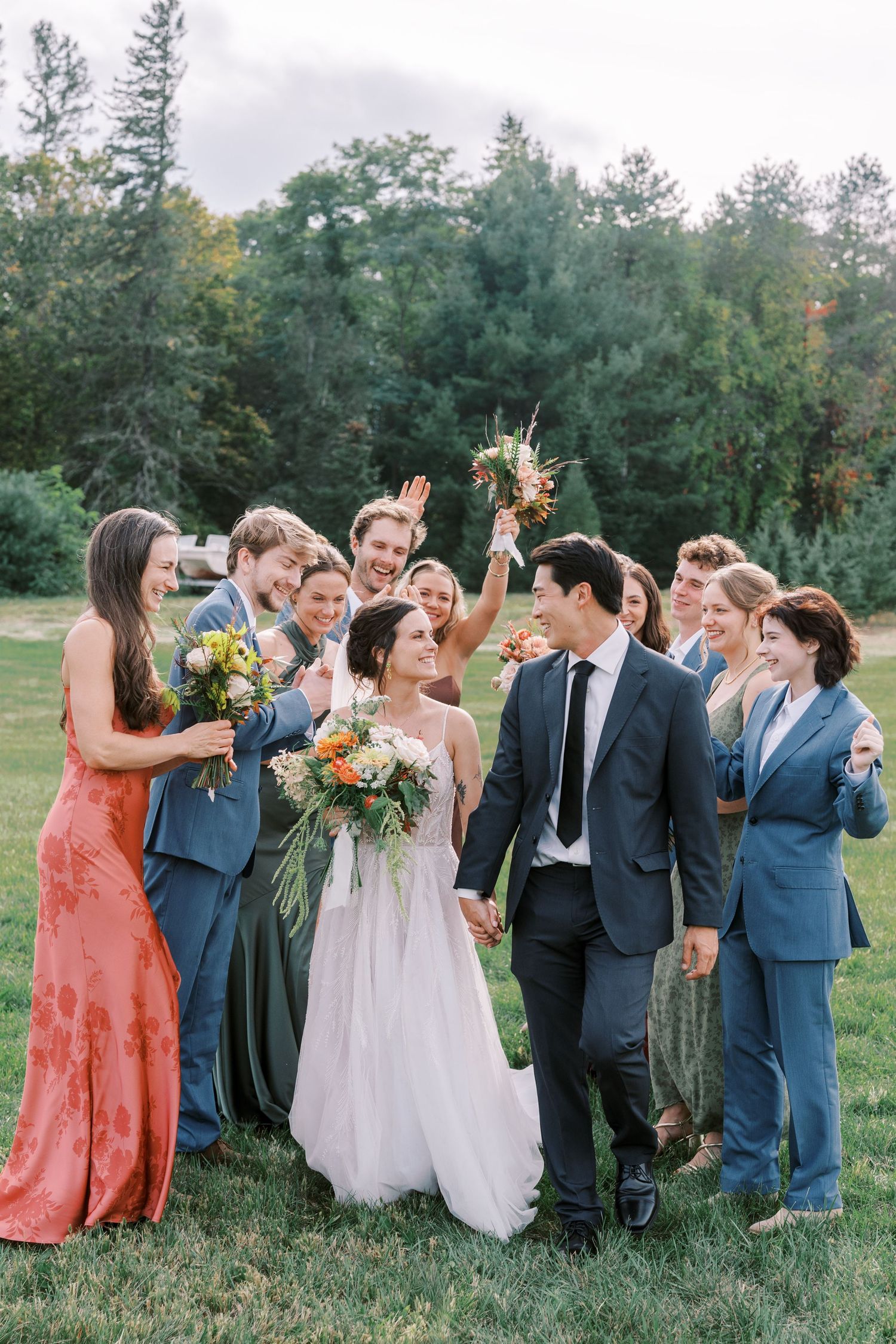Wedding party celebrates joyfully in a line on grassy field with bridesmaids in coral and sage dresses.