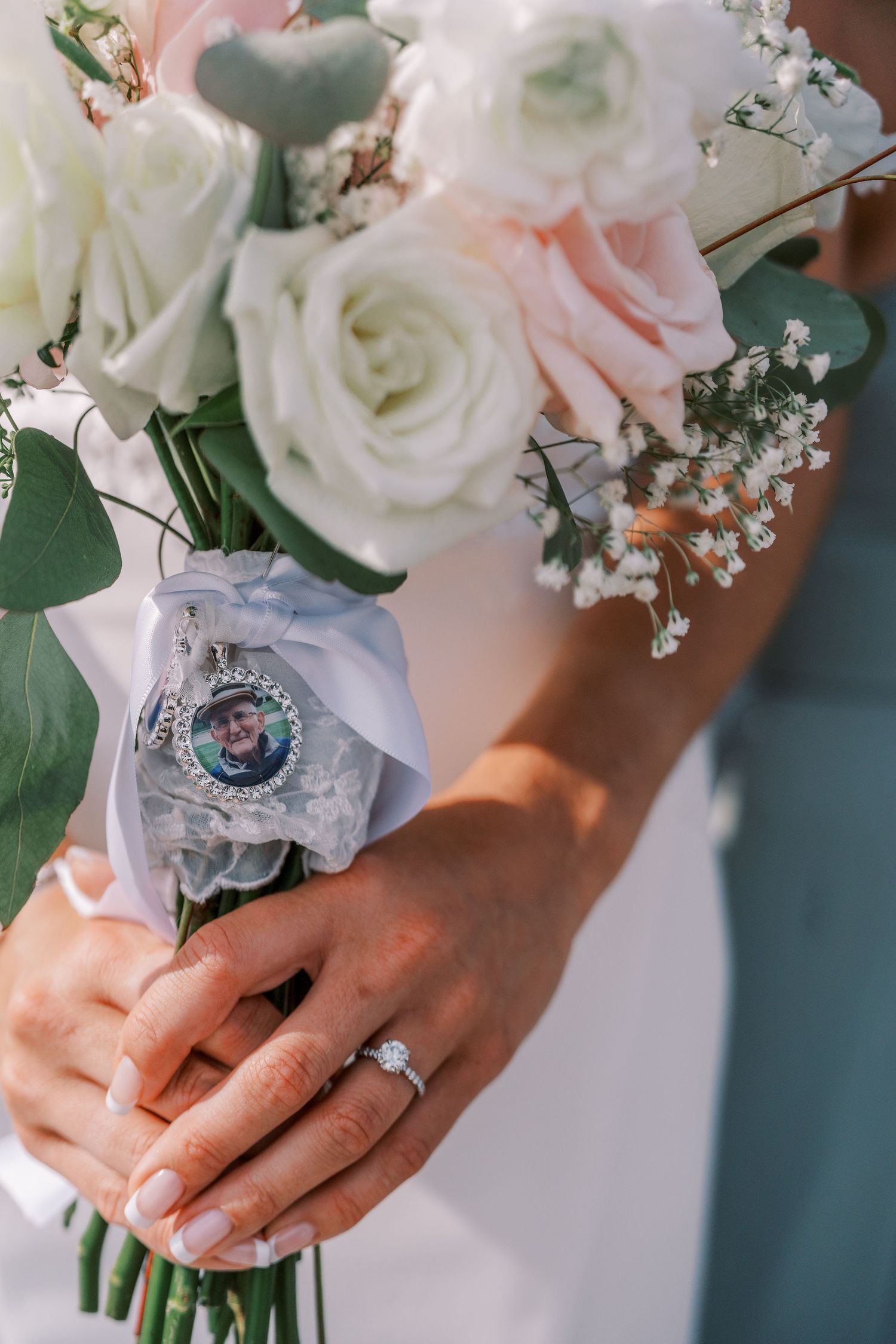 Close-up of hands holding elegant bridal bouquet with white and pink roses, baby's breath, and sentimental photo charm.