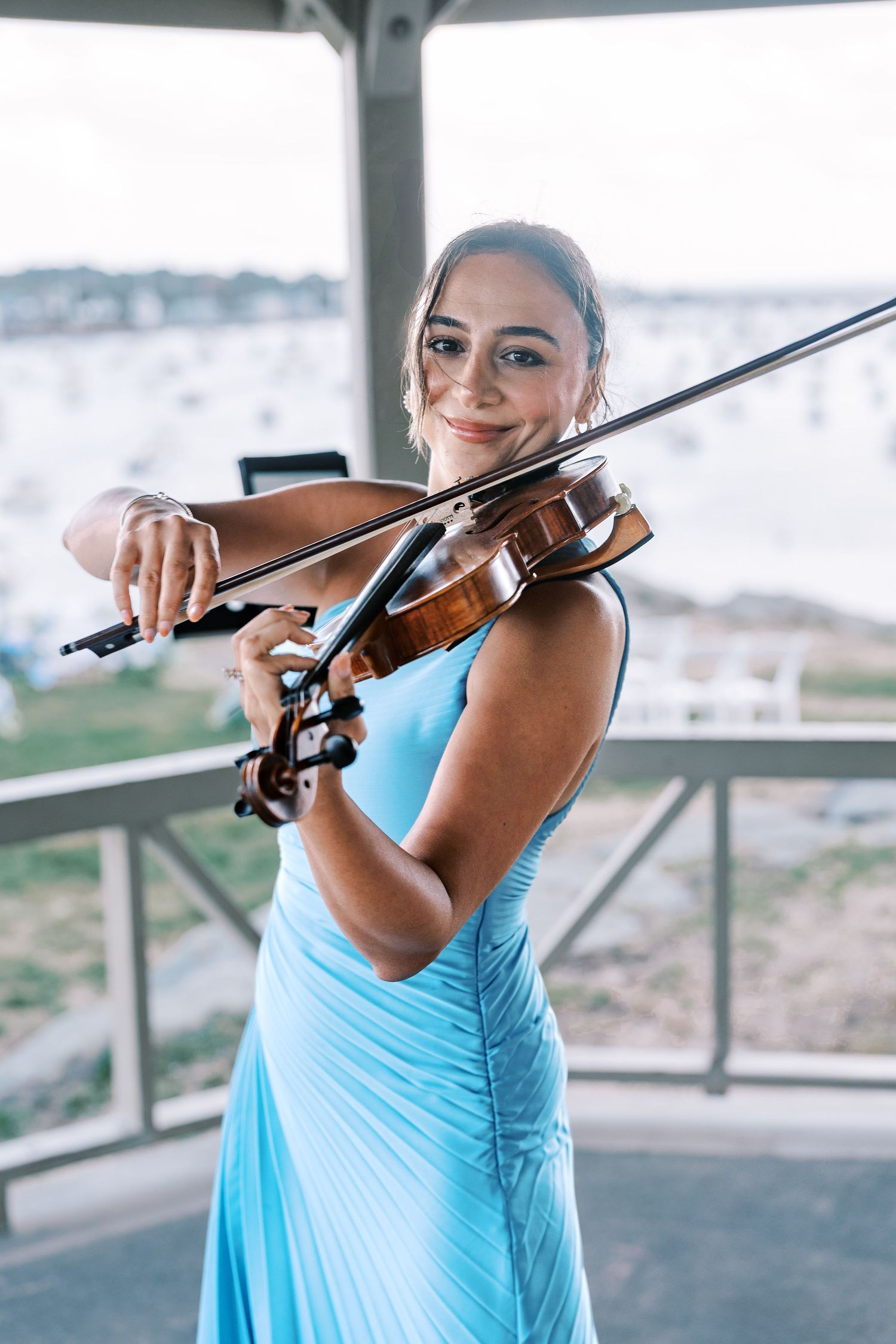 Musician in light blue dress plays violin on outdoor terrace with scenic background.