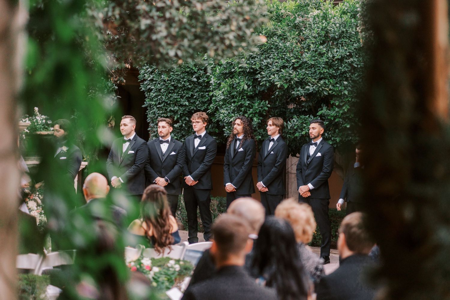 Wedding party stands in a lush garden setting during an outdoor ceremony surrounded by greenery and flowers.