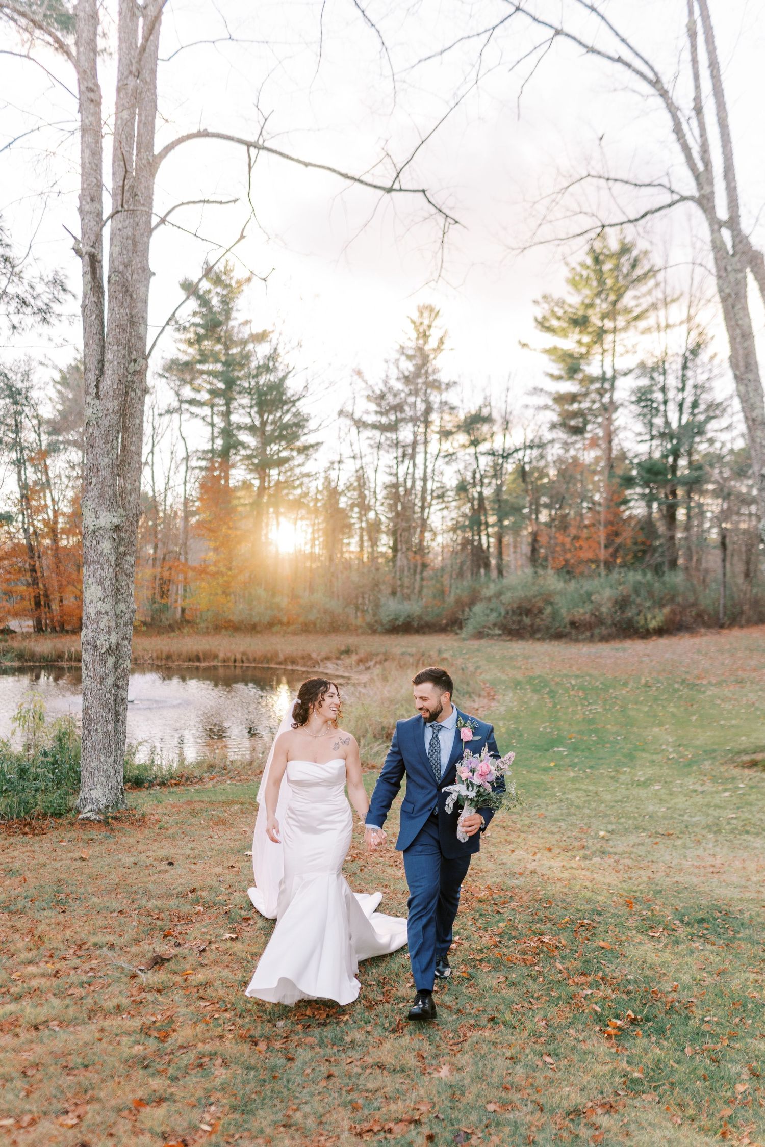 A couple walks through an autumn forest landscape with bare trees and fallen leaves at sunset.