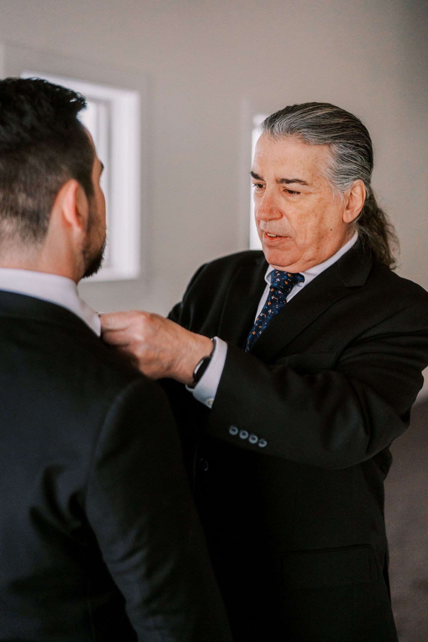 An older man in a suit adjusts another man's tie in an intimate pre-wedding moment.
