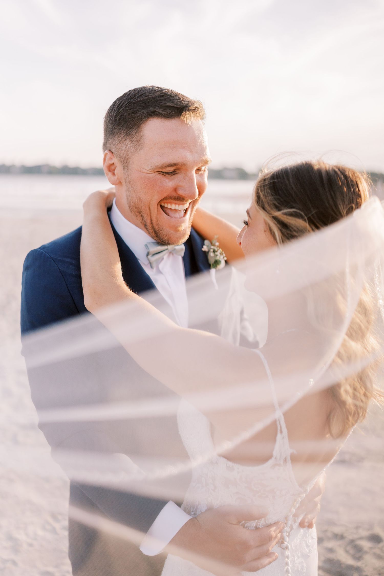 A romantic wedding couple embraces on a beach at sunset with the bride's veil flowing in the breeze.