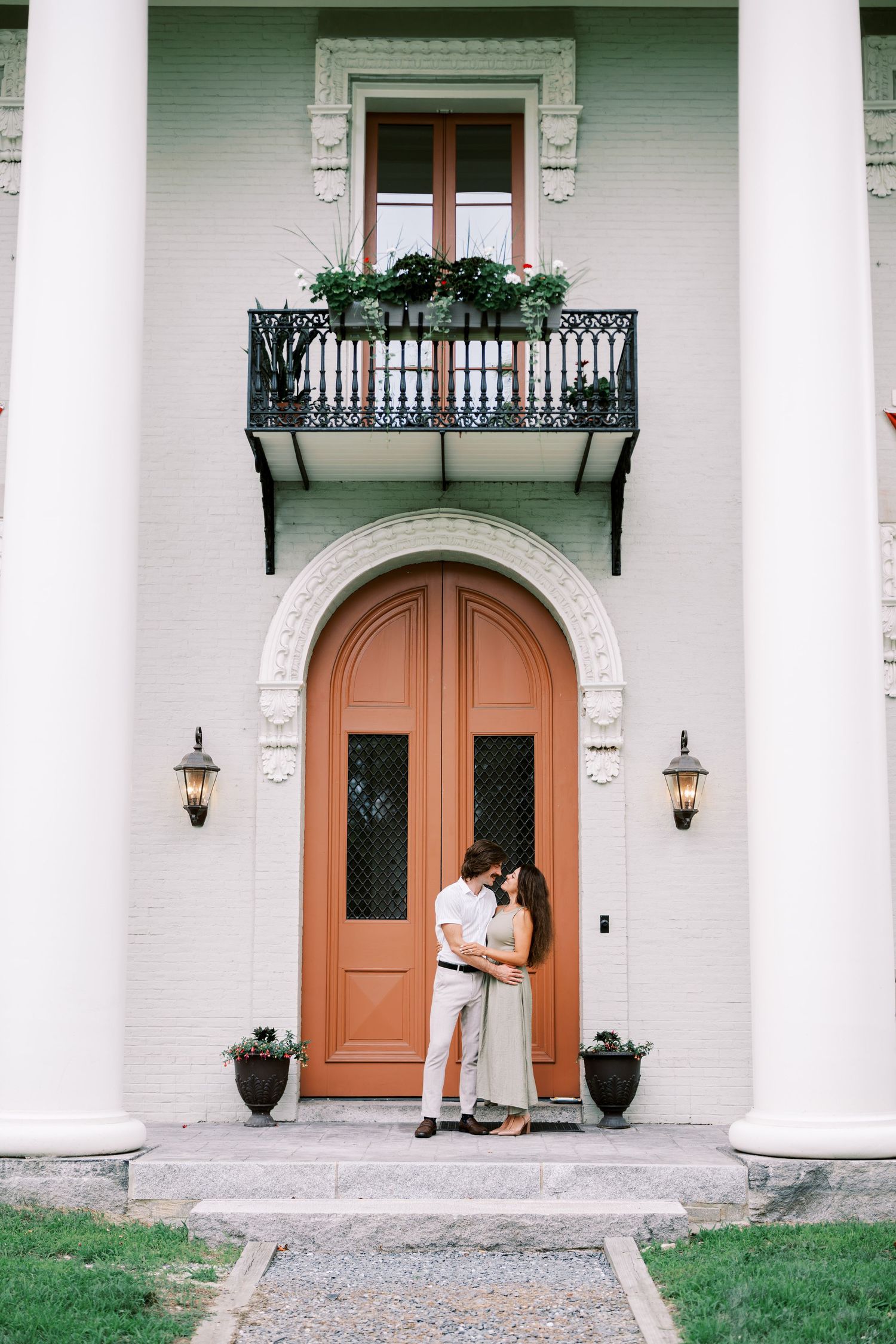 Romantic couple embracing in front of a grand white building with ornate columns and coral-colored door.