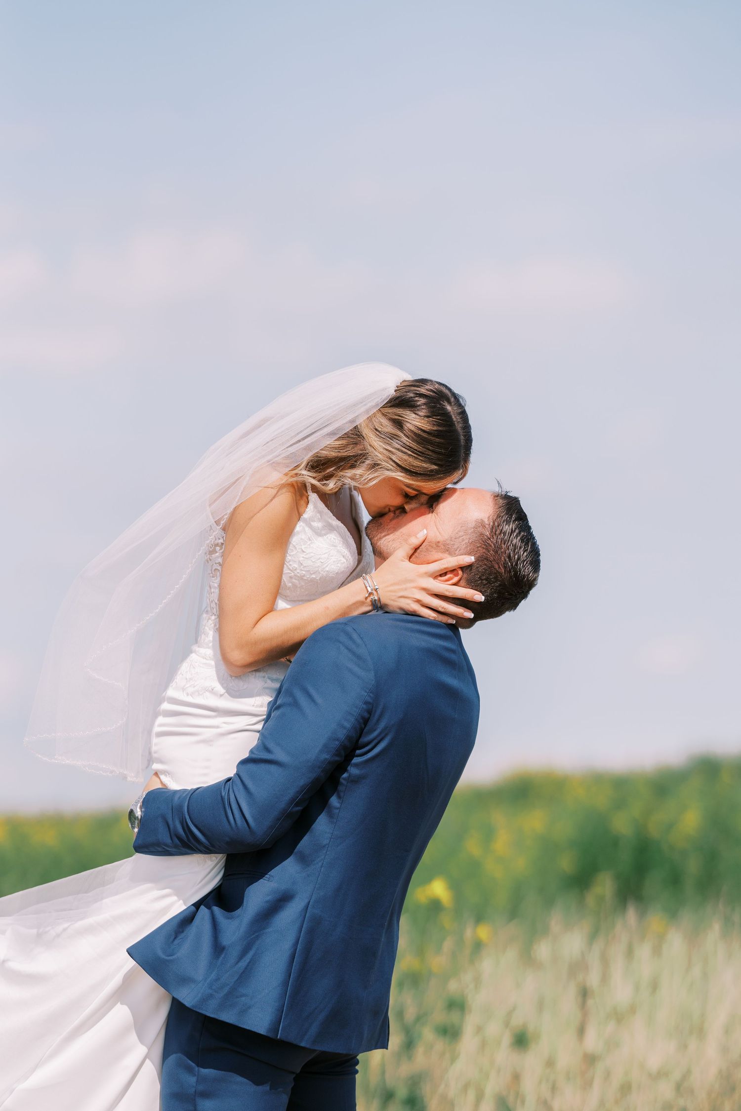 A couple in wedding attire embrace and kiss in a scenic field with tall grass at sunset.
