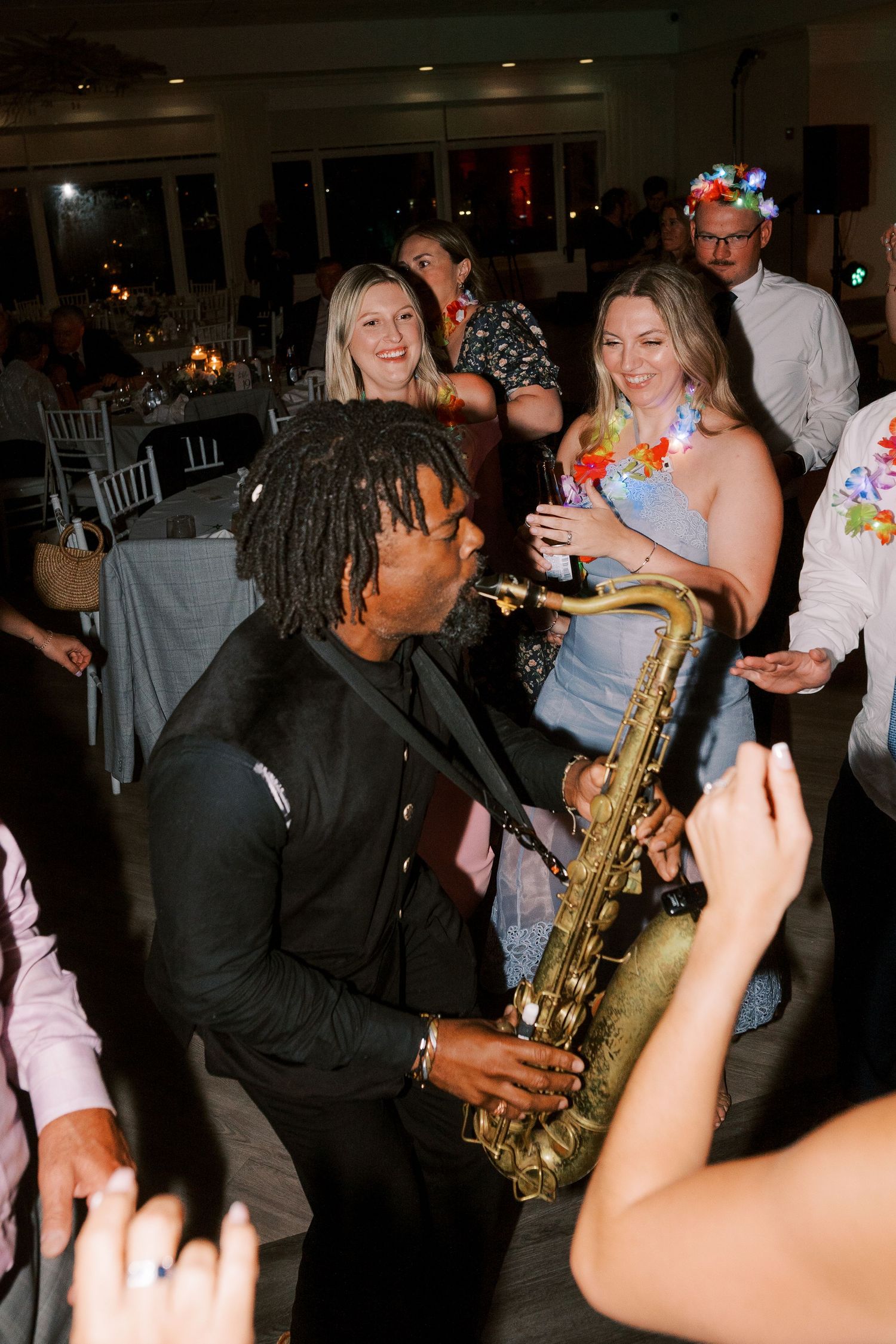 A musician in a black suit plays saxophone while interacting with guests on a lively dance floor at a party.