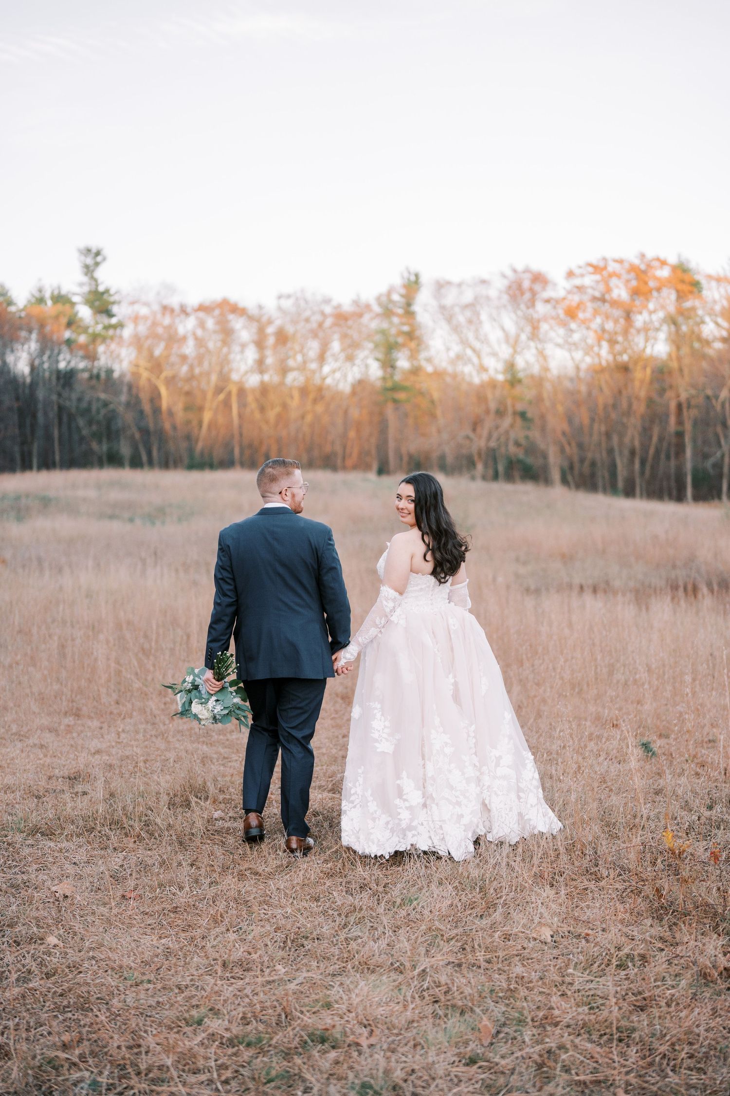 A couple walks hand in hand through a golden autumn field wearing wedding attire as the sun sets behind bare trees.