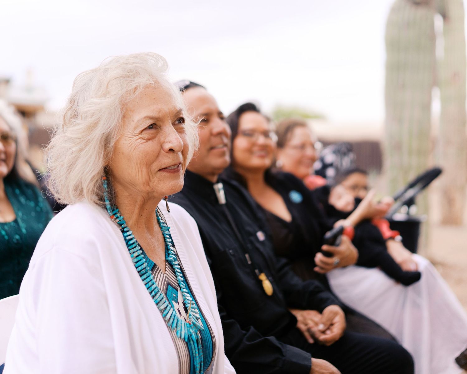 Attendees at a cultural ceremony wearing traditional turquoise jewelry and formal attire sit together outdoors.