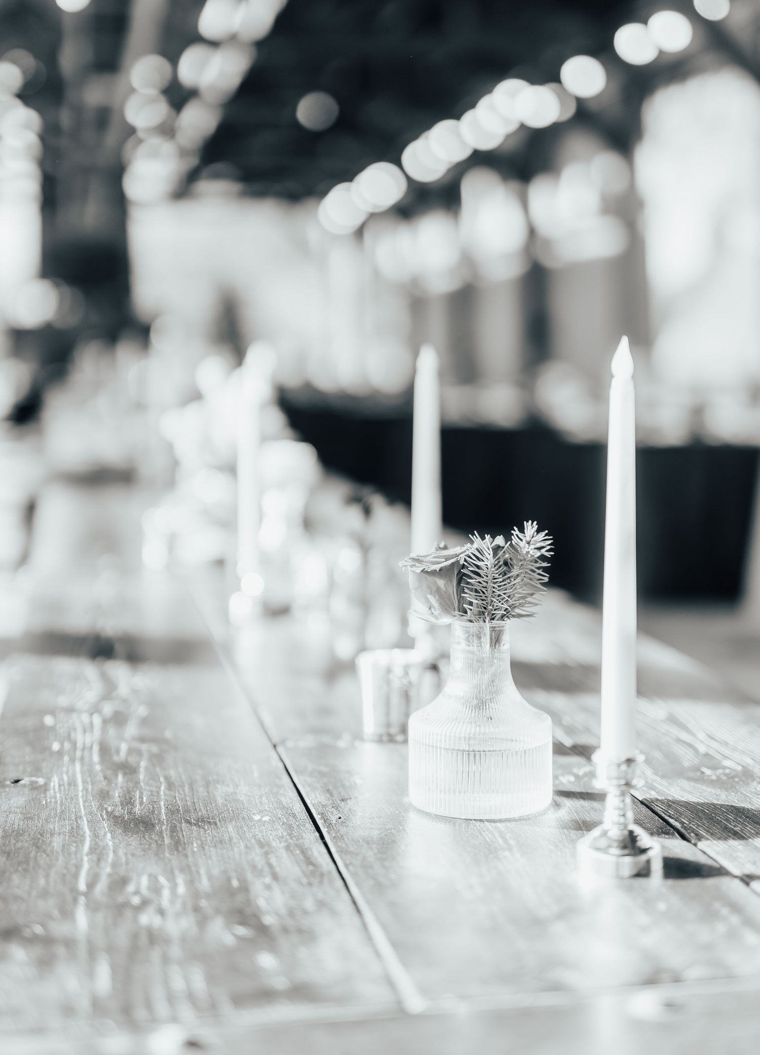White candles and small glass vase with delicate flowers on rustic wooden table create elegant table setting.