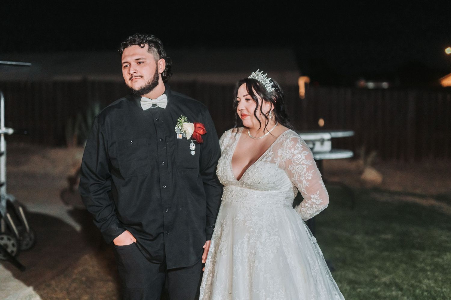 A bride and groom stand together at a nighttime outdoor wedding, with the bride wearing a lace long-sleeved gown.