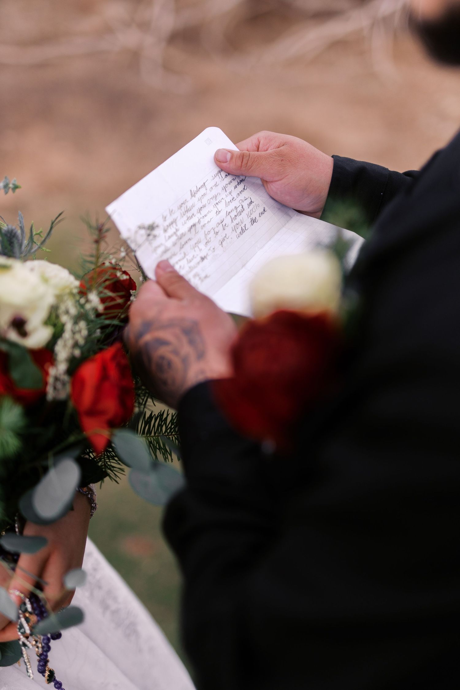A hand holds handwritten wedding vows with red and white floral arrangements visible in the background.