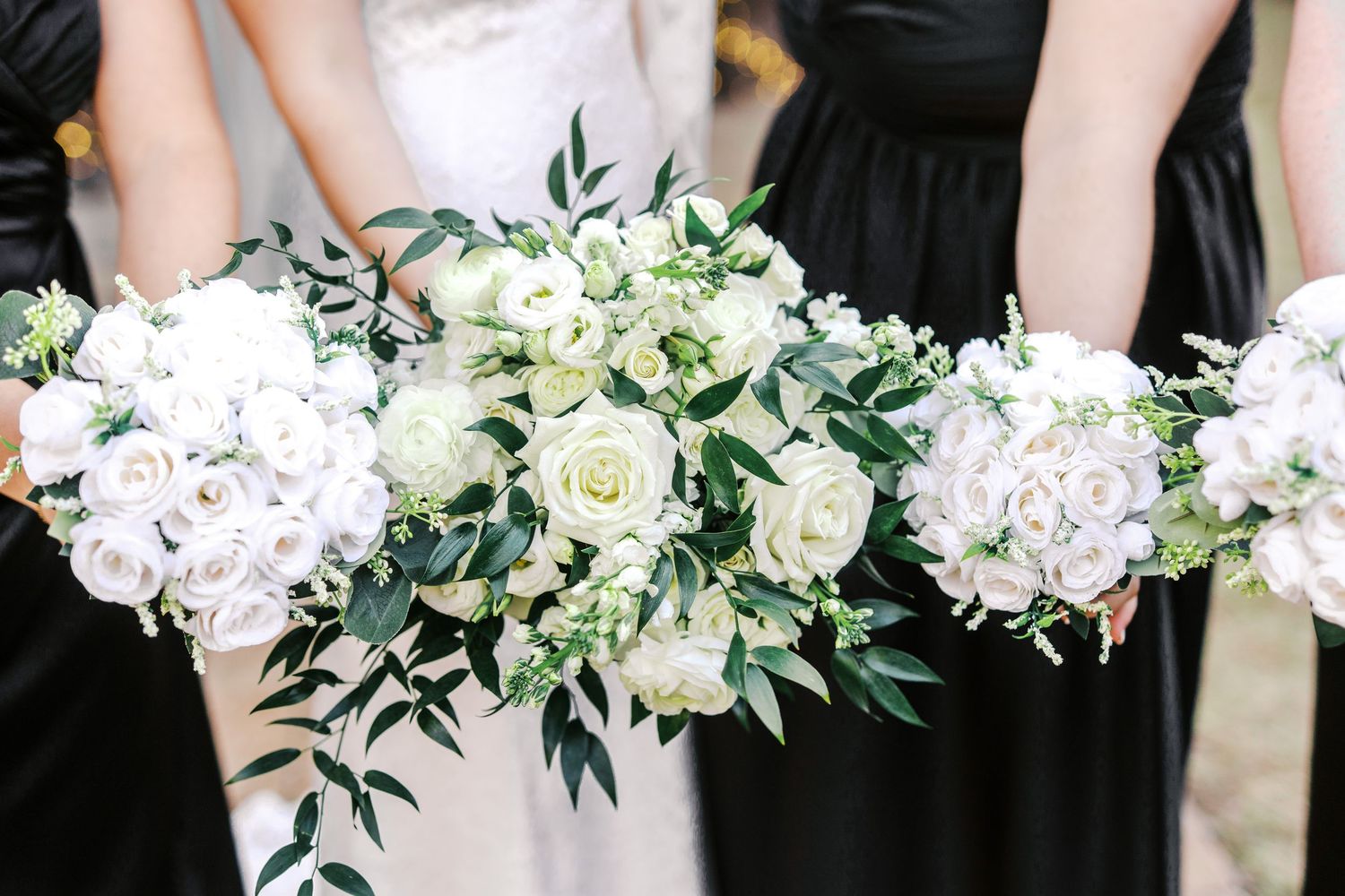 Elegant wedding bouquets featuring white hydrangeas, roses and greenery held against black bridesmaid dresses.