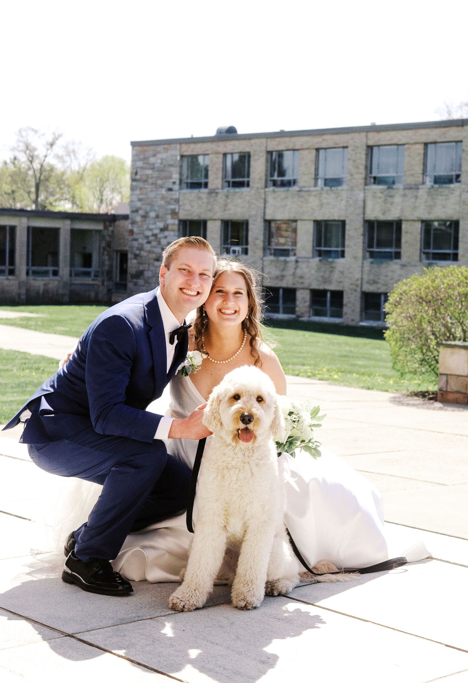 A couple poses with their fluffy white Goldendoodle dog on their wedding day outside a stone building.