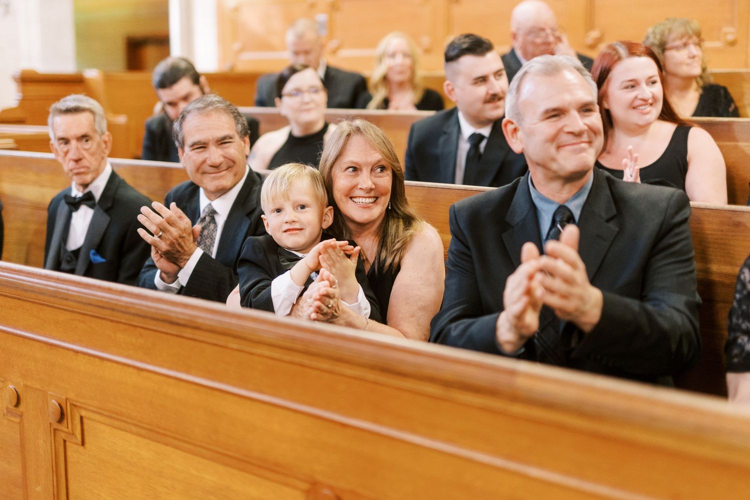Wedding guests seated in wooden church pews smile and applaud during a ceremony.