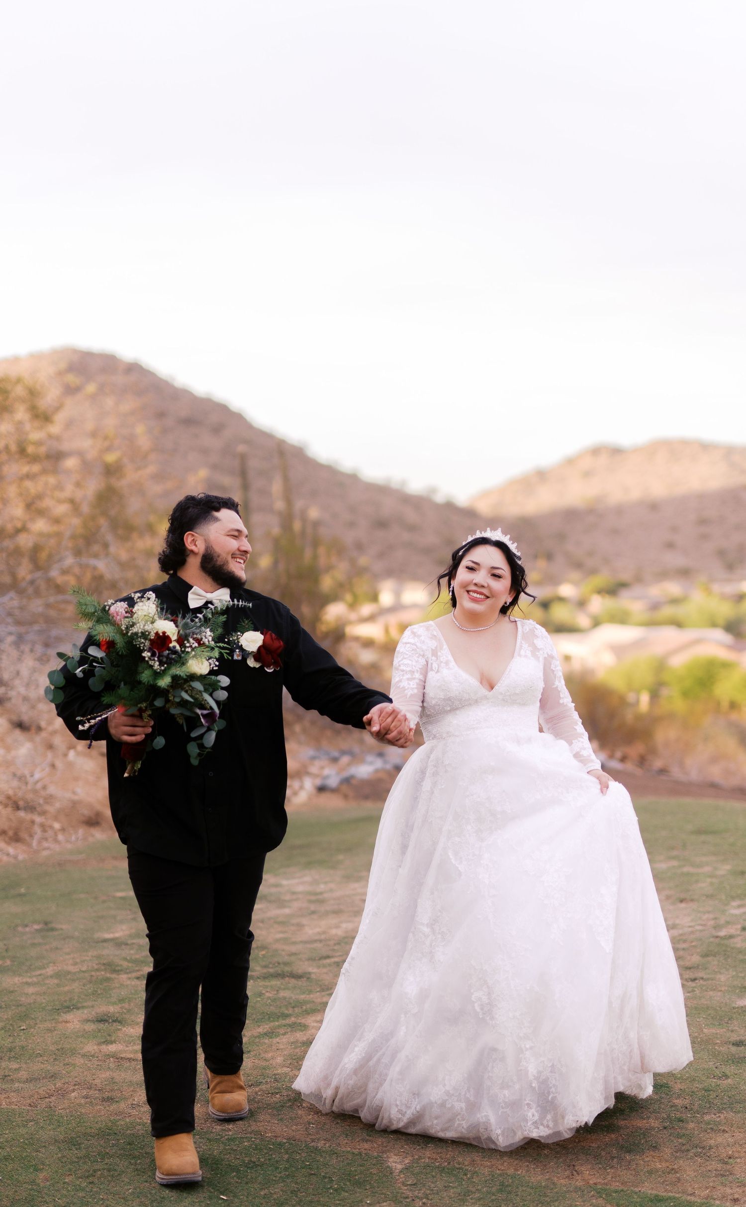 Two people in wedding attire run joyfully across desert landscape holding hands at sunset.