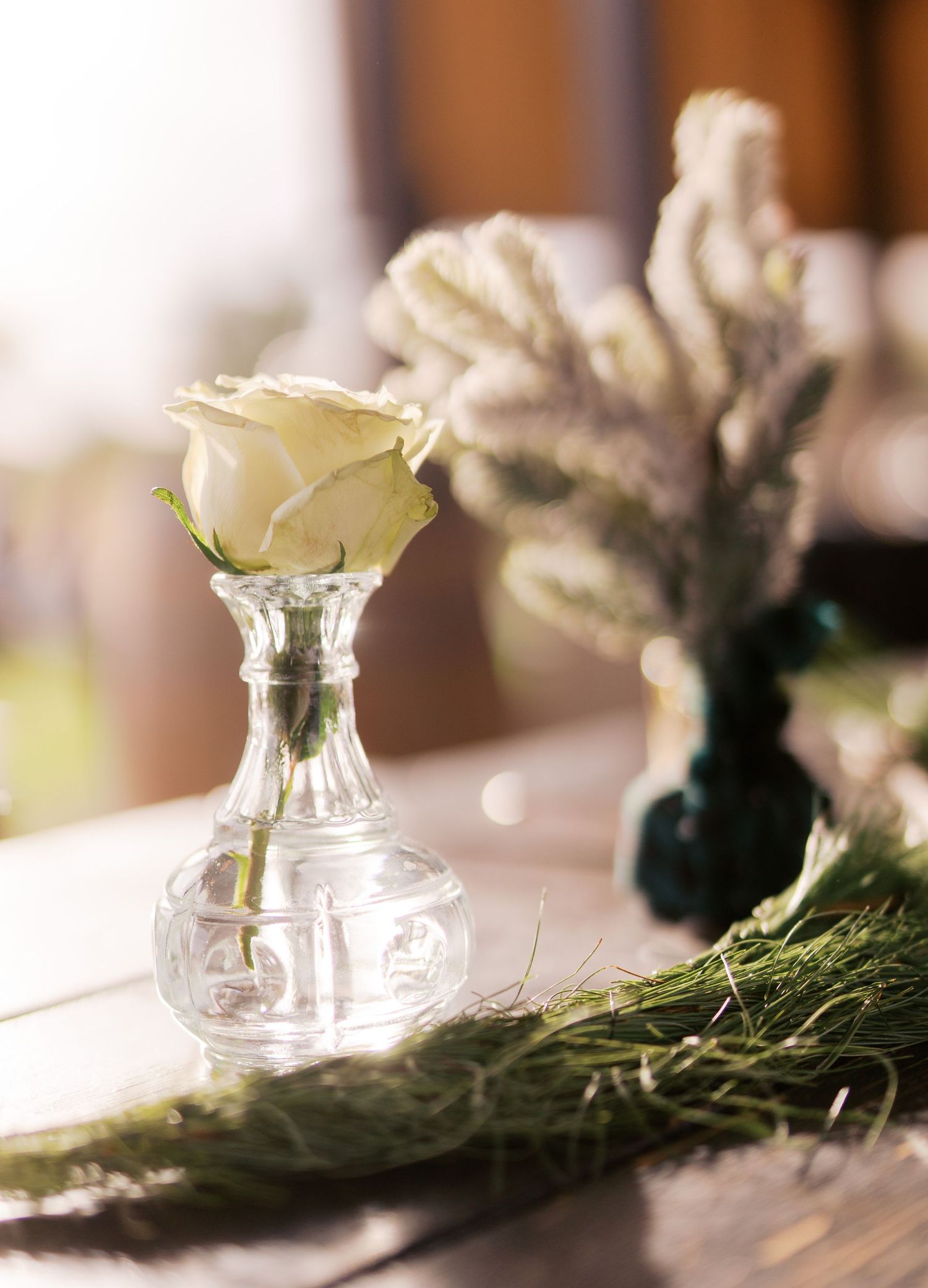 A single white rose in a crystal vase sits atop a table with delicate greenery and rustic decor elements.