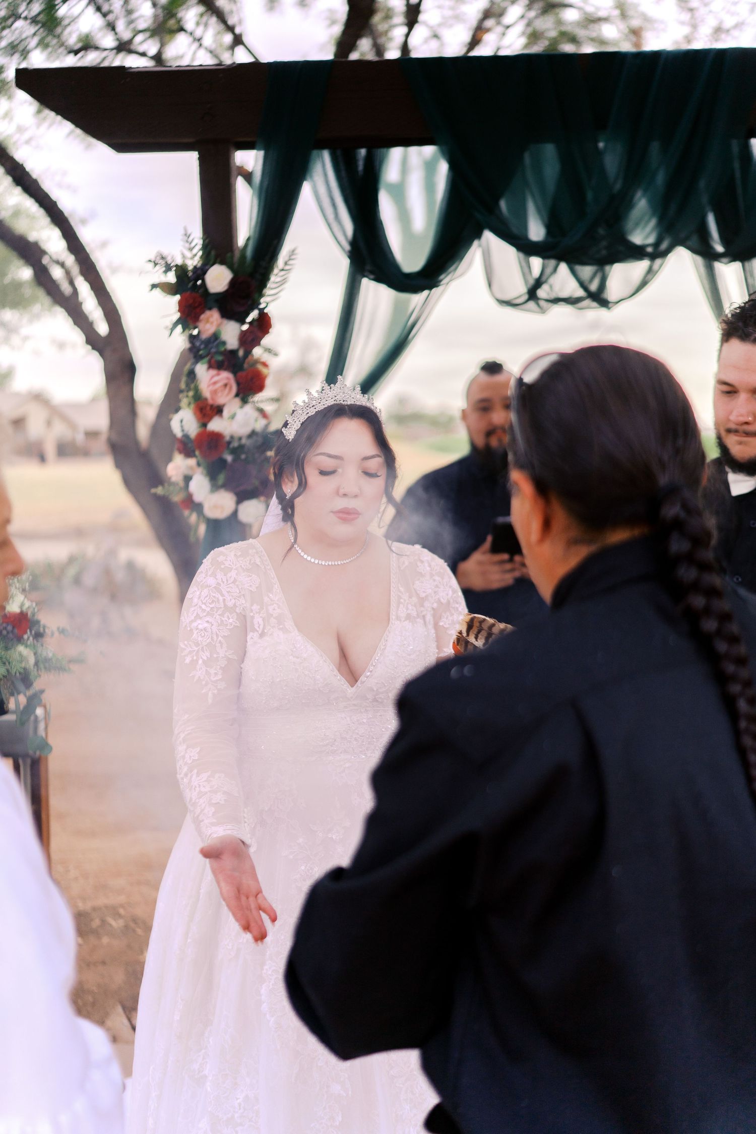 Romantic outdoor wedding ceremony under a wooden arch decorated with dark teal fabric and floral arrangements.