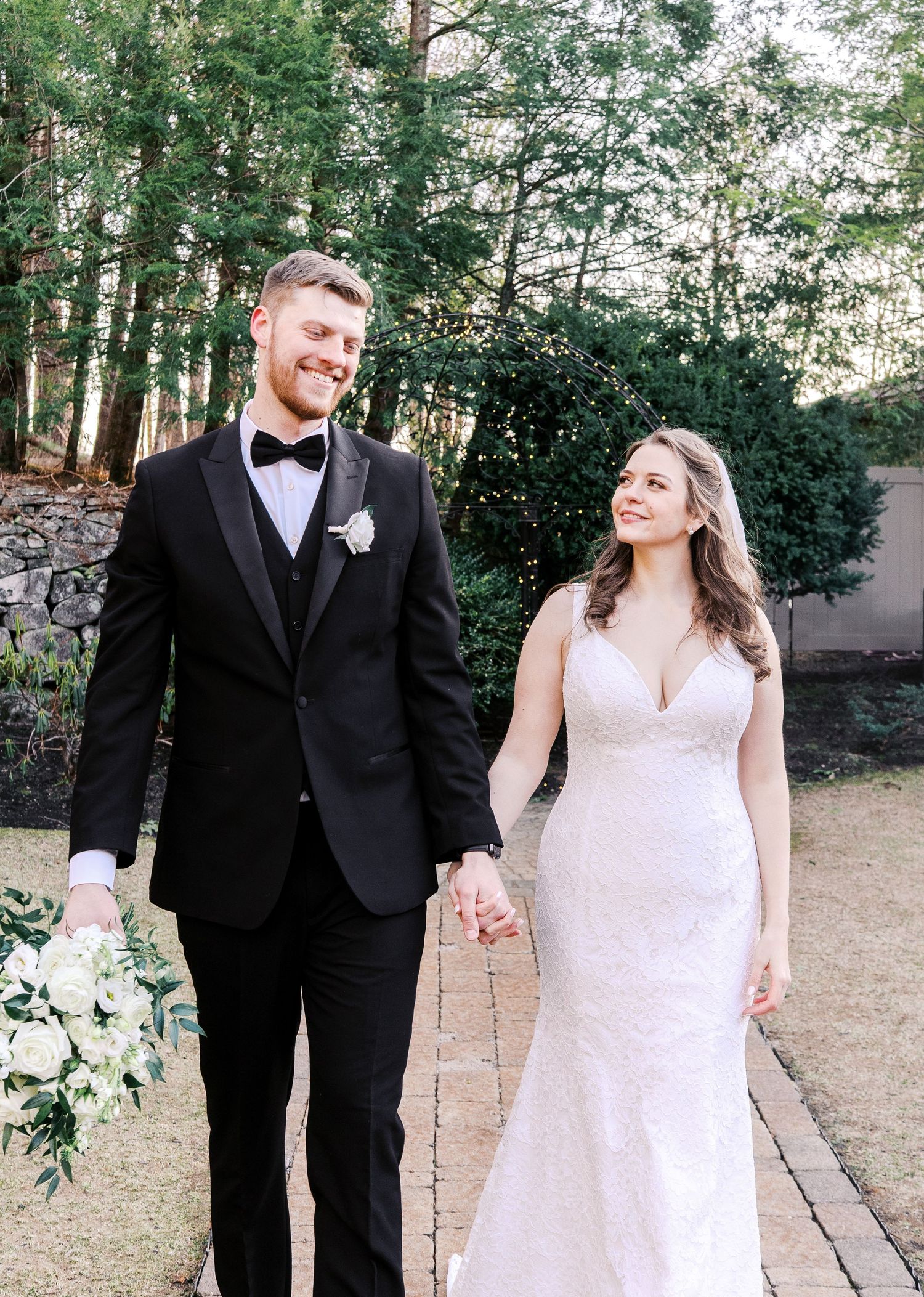 Newlyweds walking together outdoors along a stone path surrounded by trees.