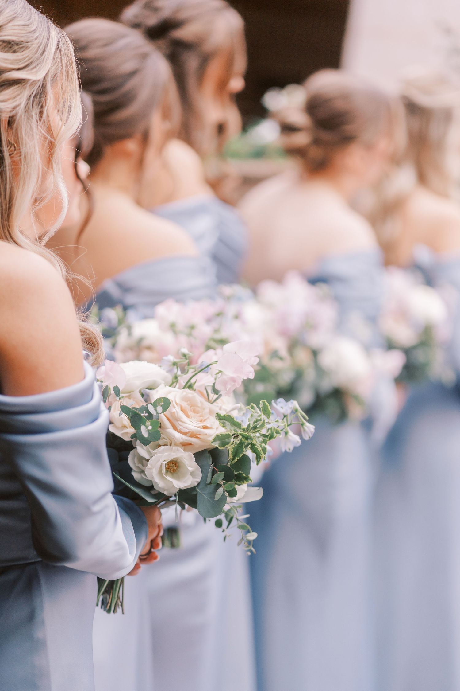 Bridesmaids in dusty blue dresses stand in line holding delicate pink and white floral bouquets at a wedding ceremony.