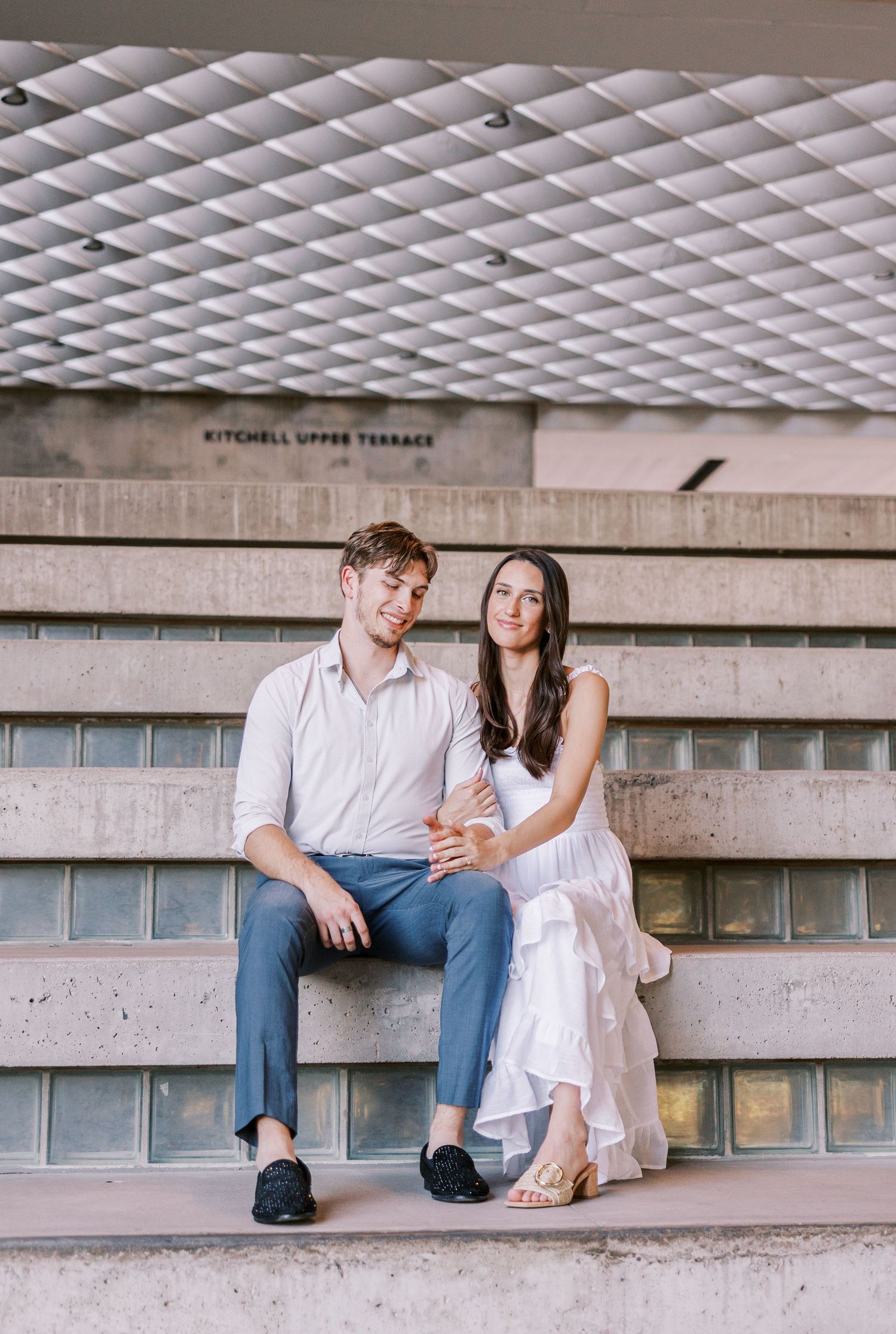 Couple sitting close together on concrete steps under geometric ceiling.