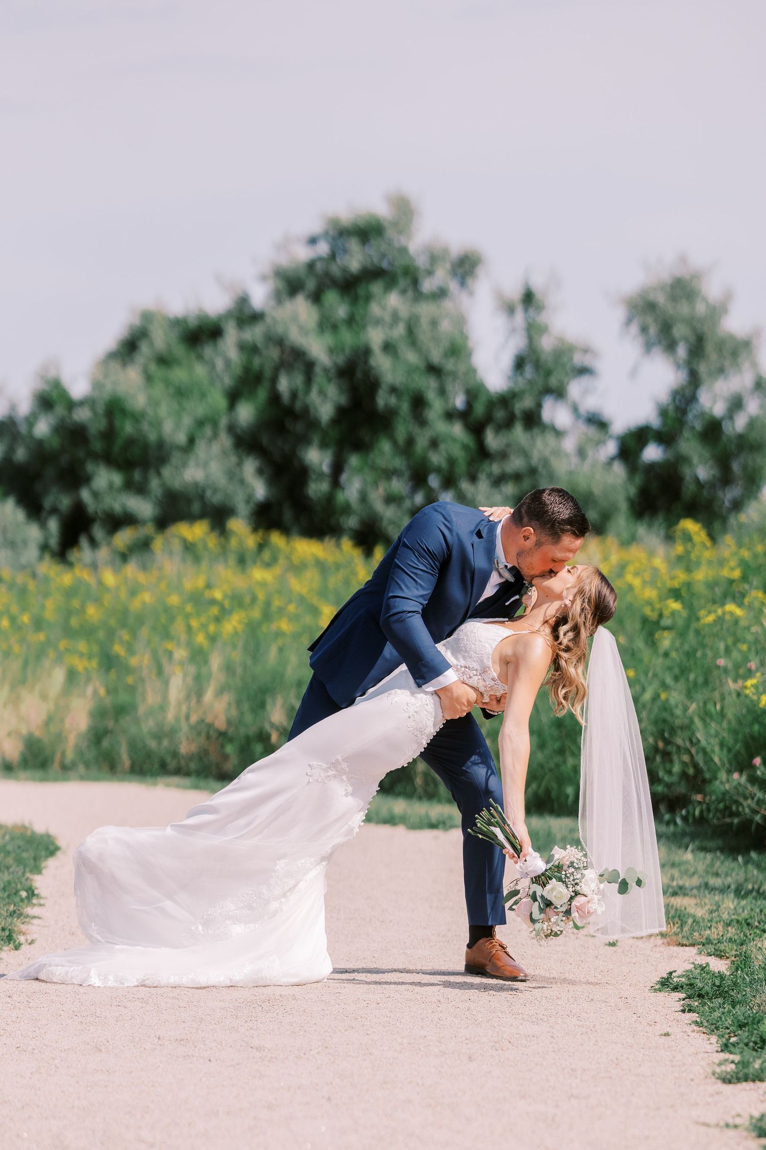 A romantic wedding couple shares a dip kiss in a field of yellow wildflowers on a sunny day.