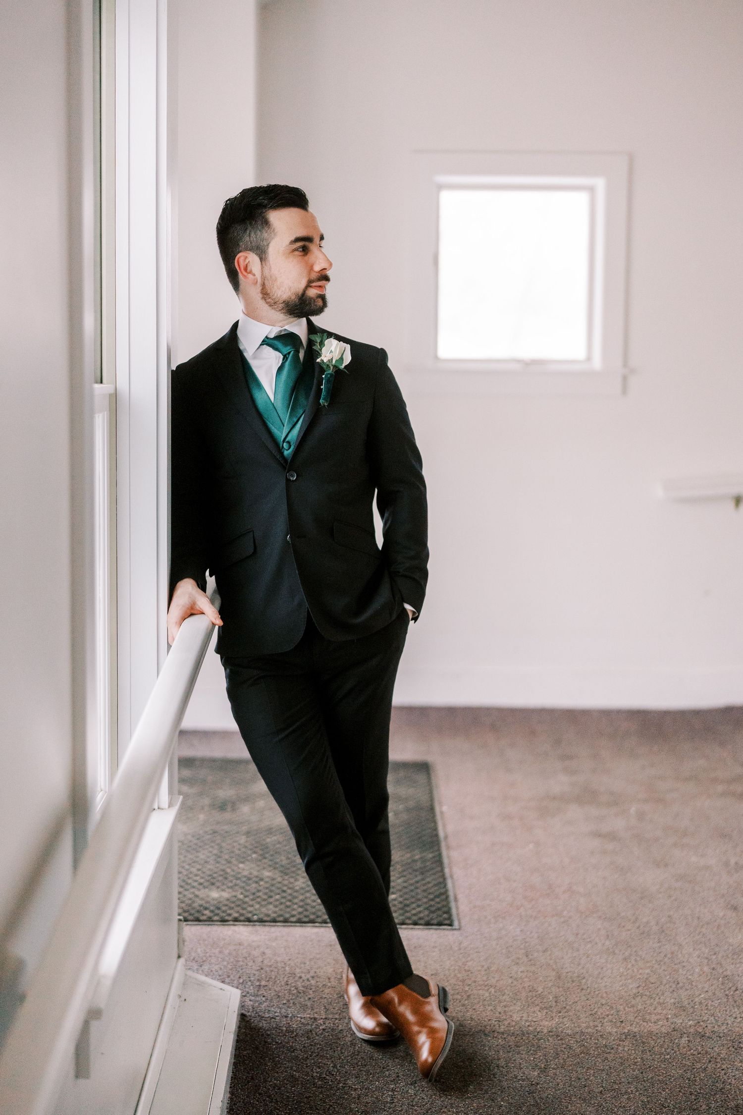 A person in a black suit with teal tie poses confidently against a white wall.