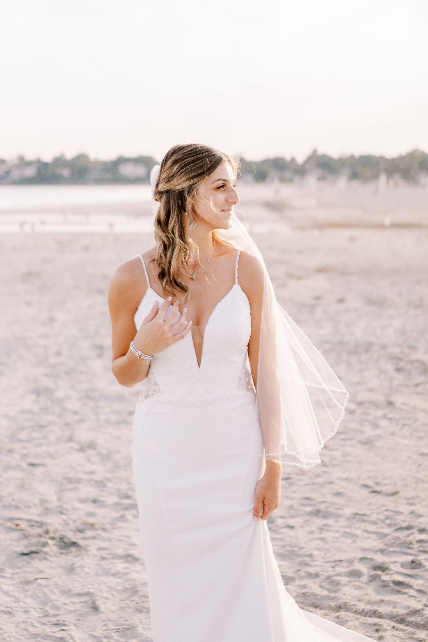 A bride in a flowing white dress laughs joyfully while standing on a sandy beach at sunset.