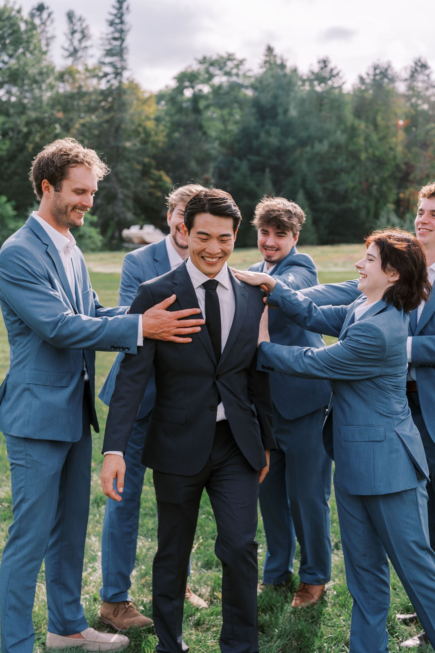 Groomsmen in blue suits share a laugh with groom in black suit during outdoor wedding photos.