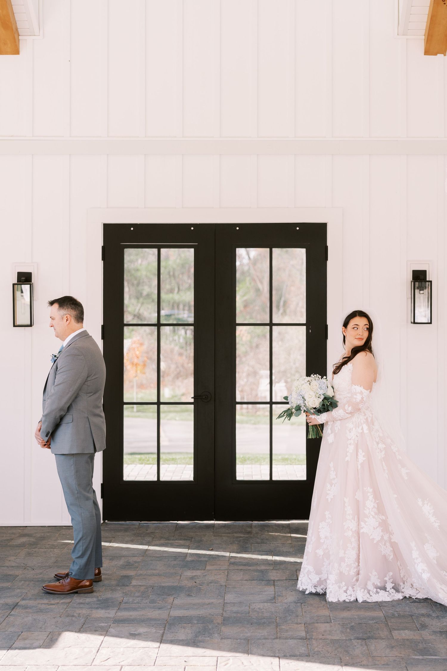 A bride in a lace wedding dress and groom in gray suit stand apart for a first look photo by black French doors.
