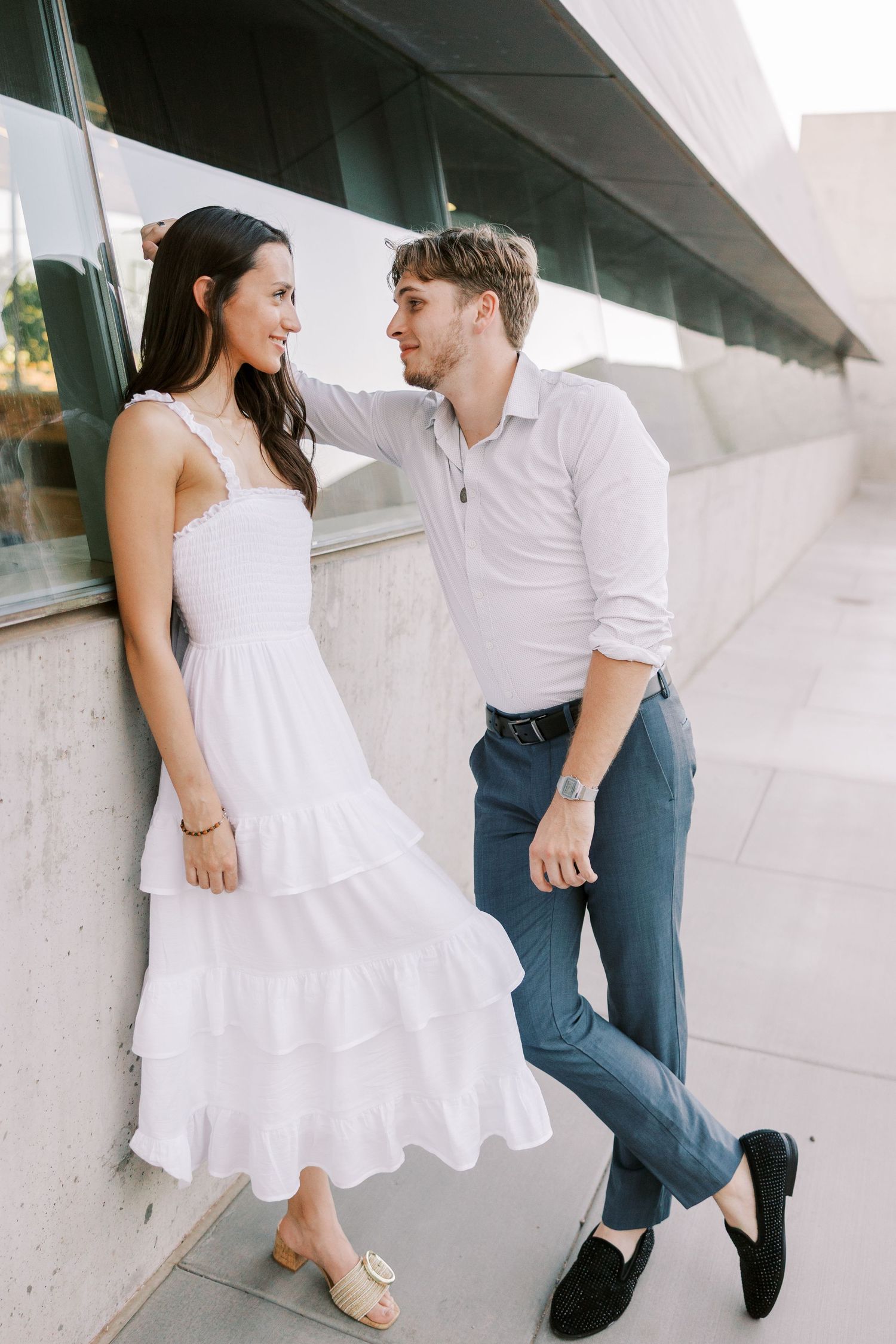 Couple in white and blue outfits playfully posing against a white building wall.