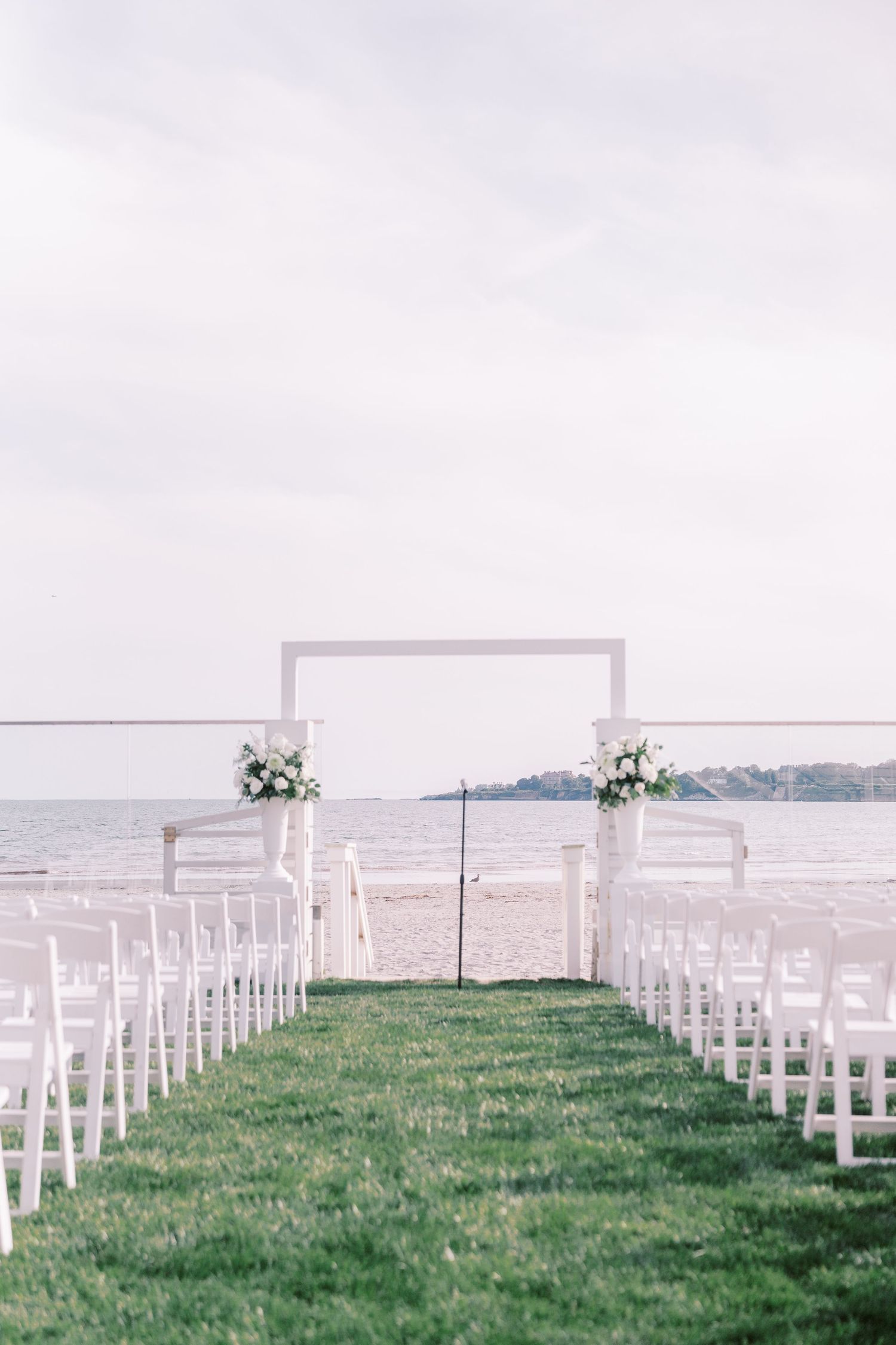 White chairs line a grassy outdoor wedding ceremony setup overlooking a waterfront view with floral arrangements at the altar.