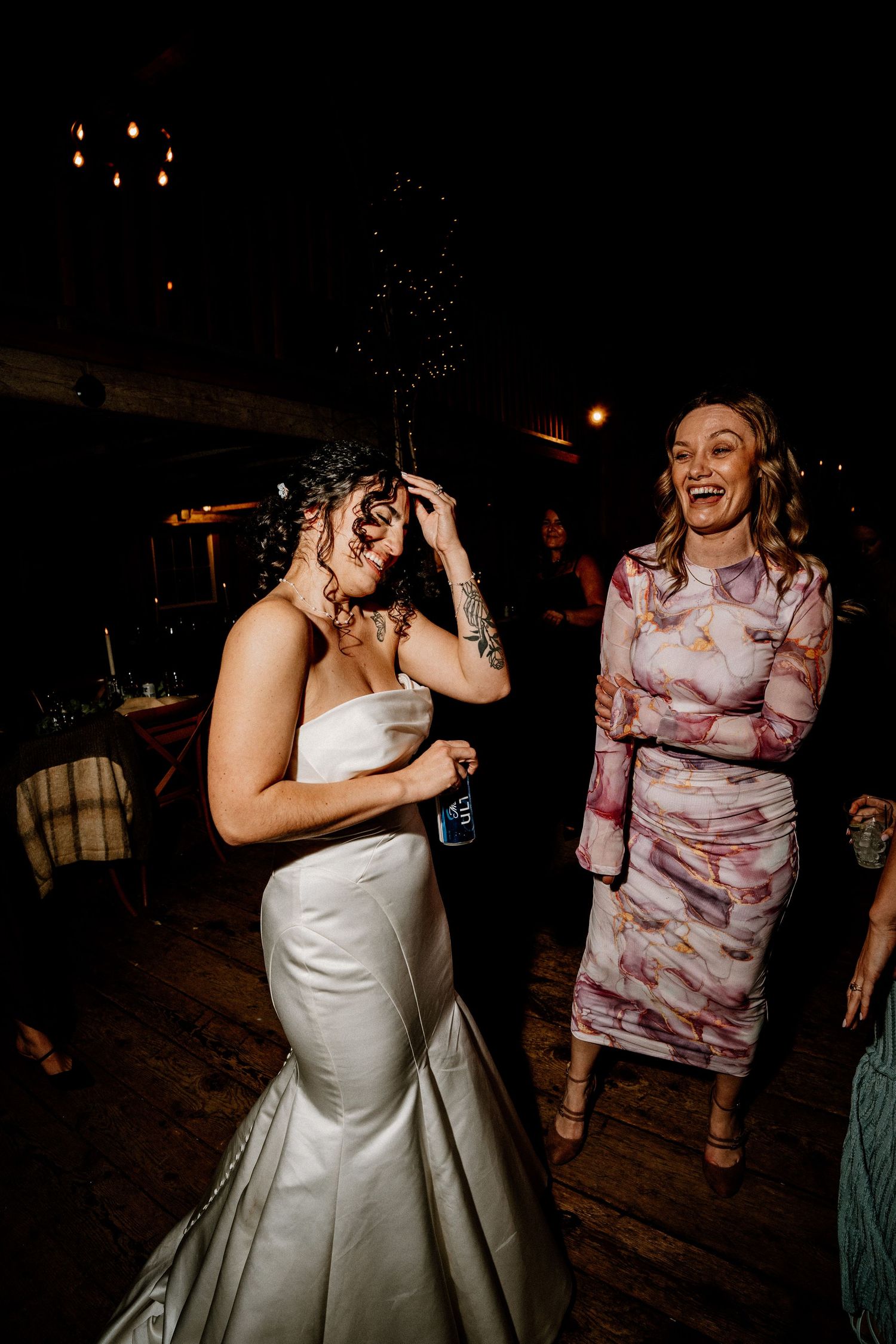 Two women share a joyful moment on a dance floor at a wedding reception, with one wearing a white gown and another in pink.
