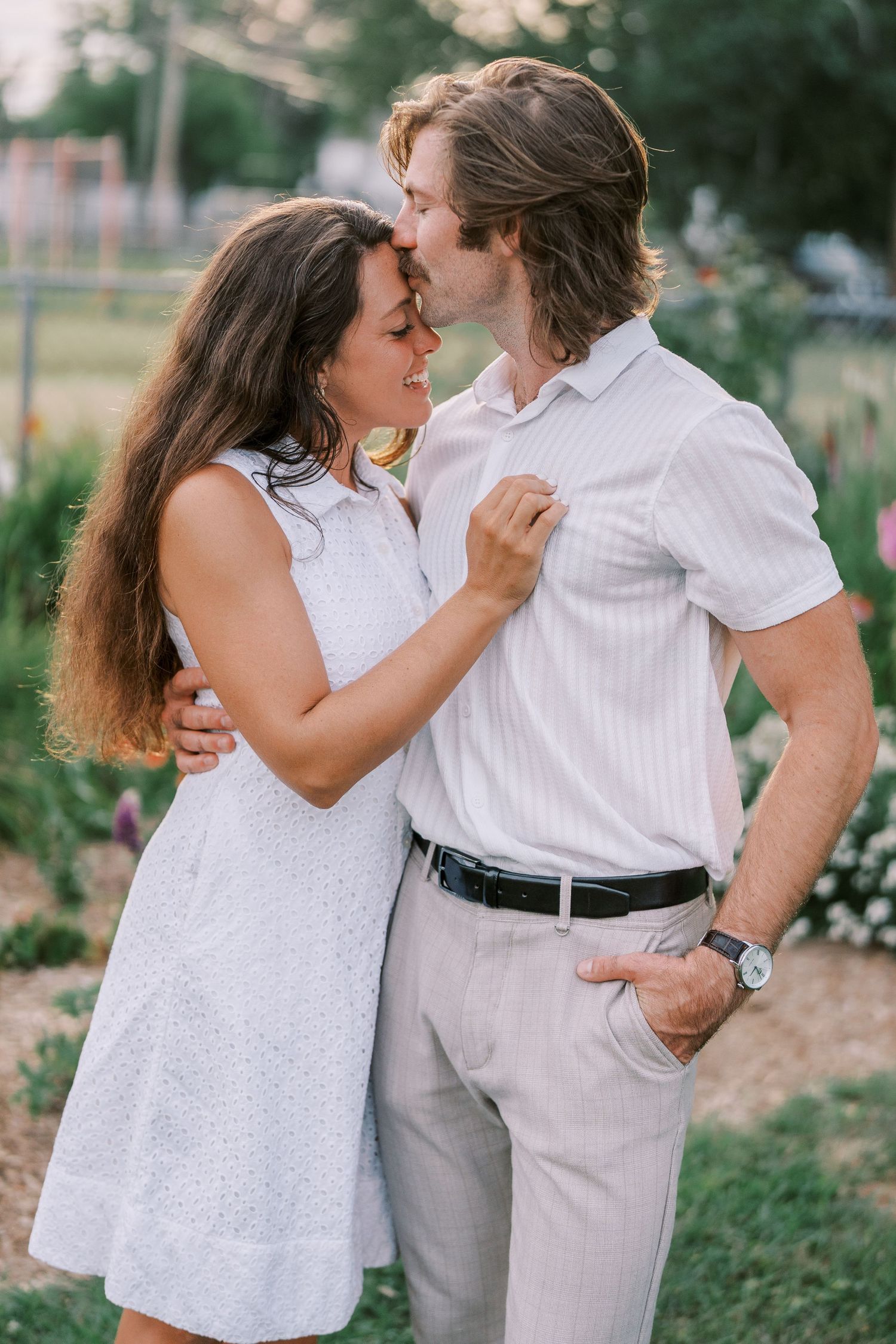 Playful couple embracing in white and grey outfits against floral backdrop.