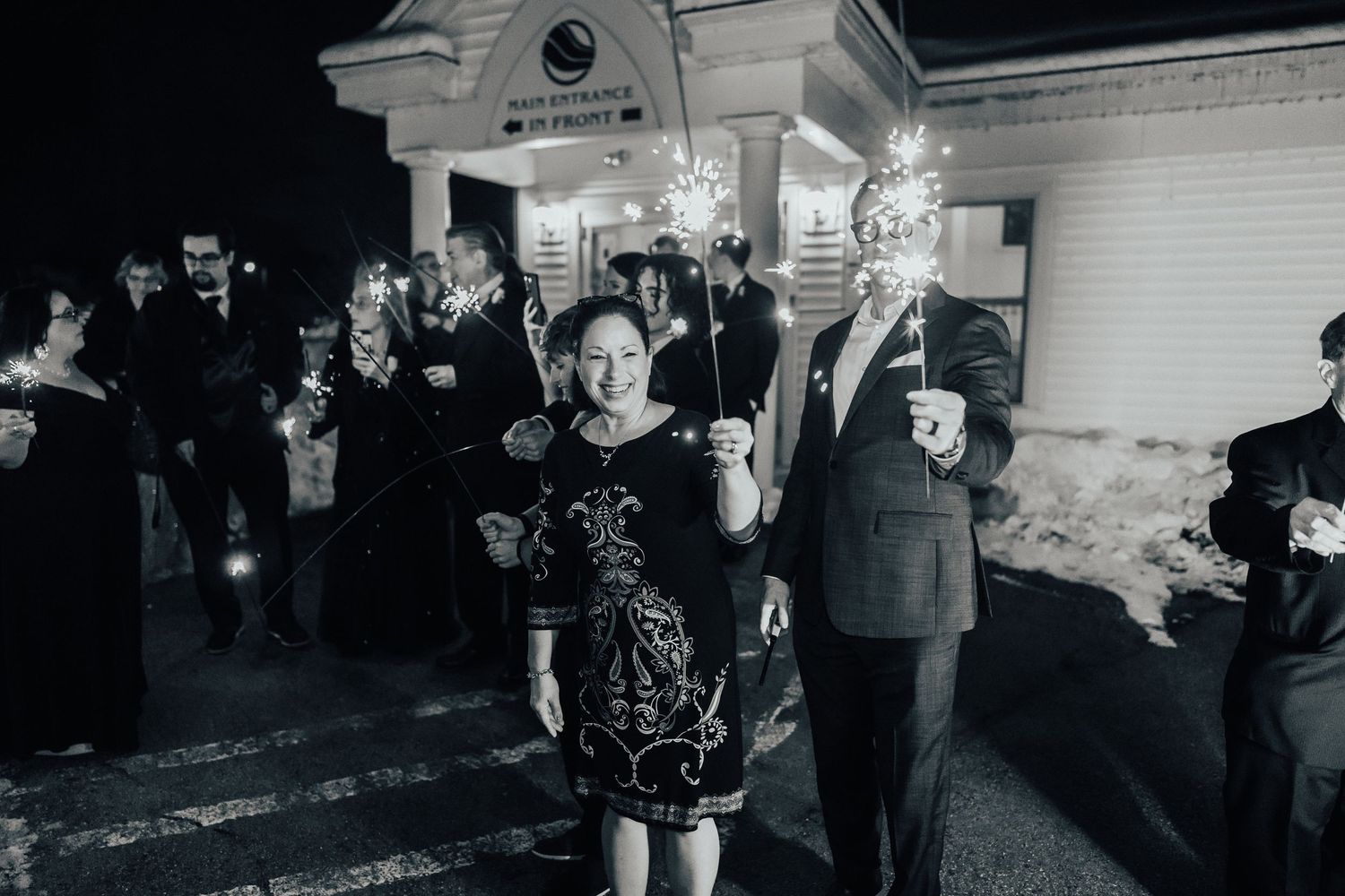 A black and white photograph shows people celebrating outside a building at night with sparklers.