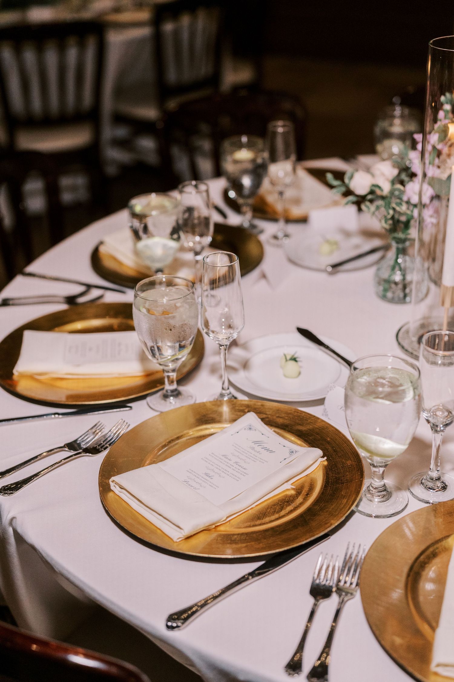 Elegant wedding table setting with gold charger plates, white linens, crystal glasses and delicate floral centerpieces.