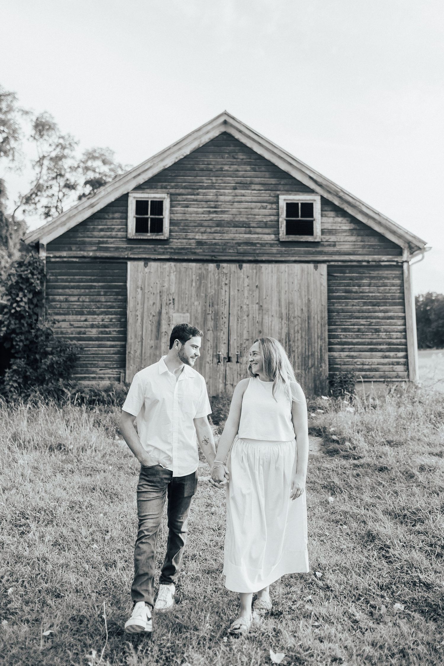 A couple poses in front of a rustic wooden barn in a grassy field during a photo session.