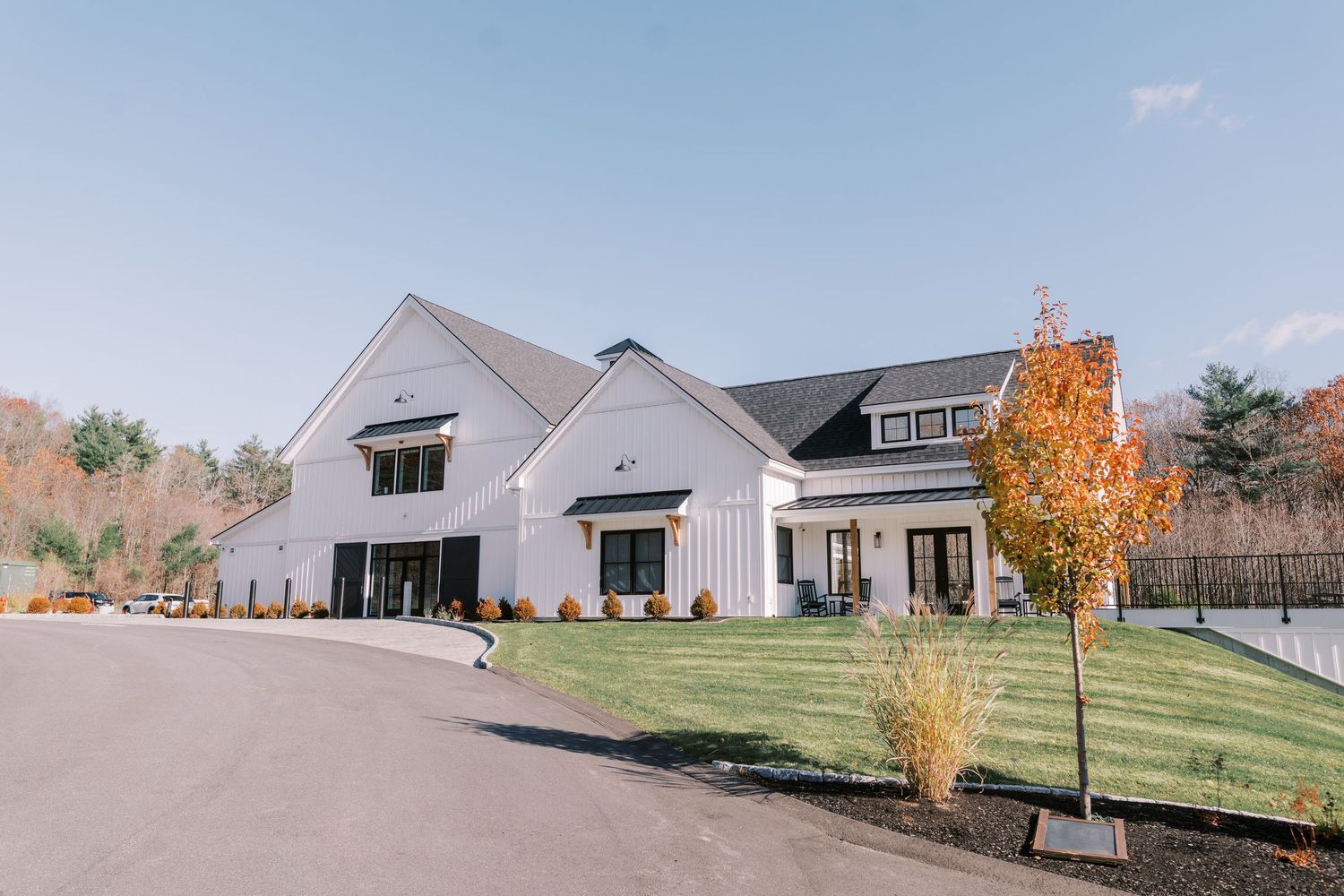 Modern white farmhouse style home with black trim features a circular driveway and autumn trees on a manicured lawn.