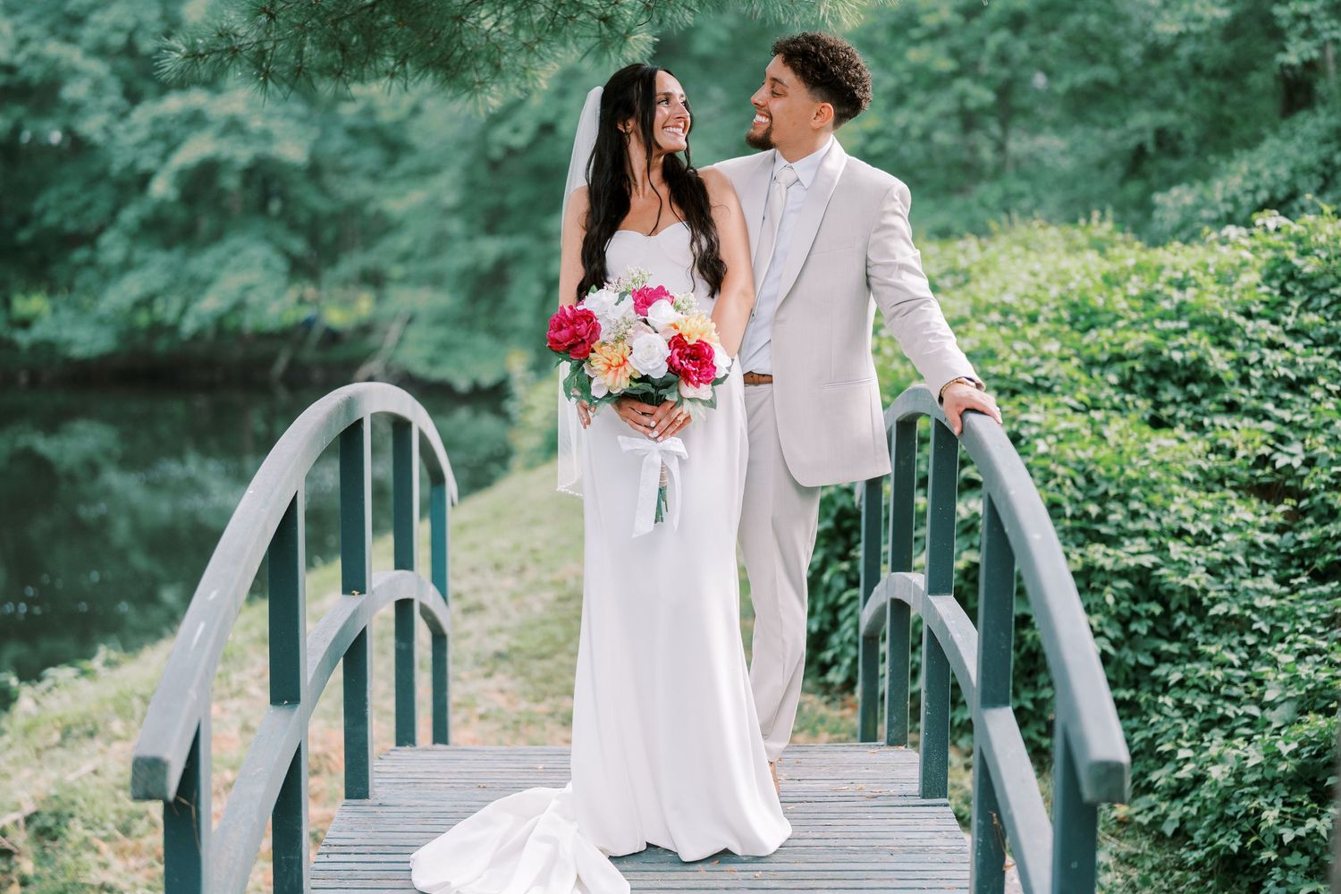 Newlyweds pose on garden bridge with pink bouquet and light gray suit.