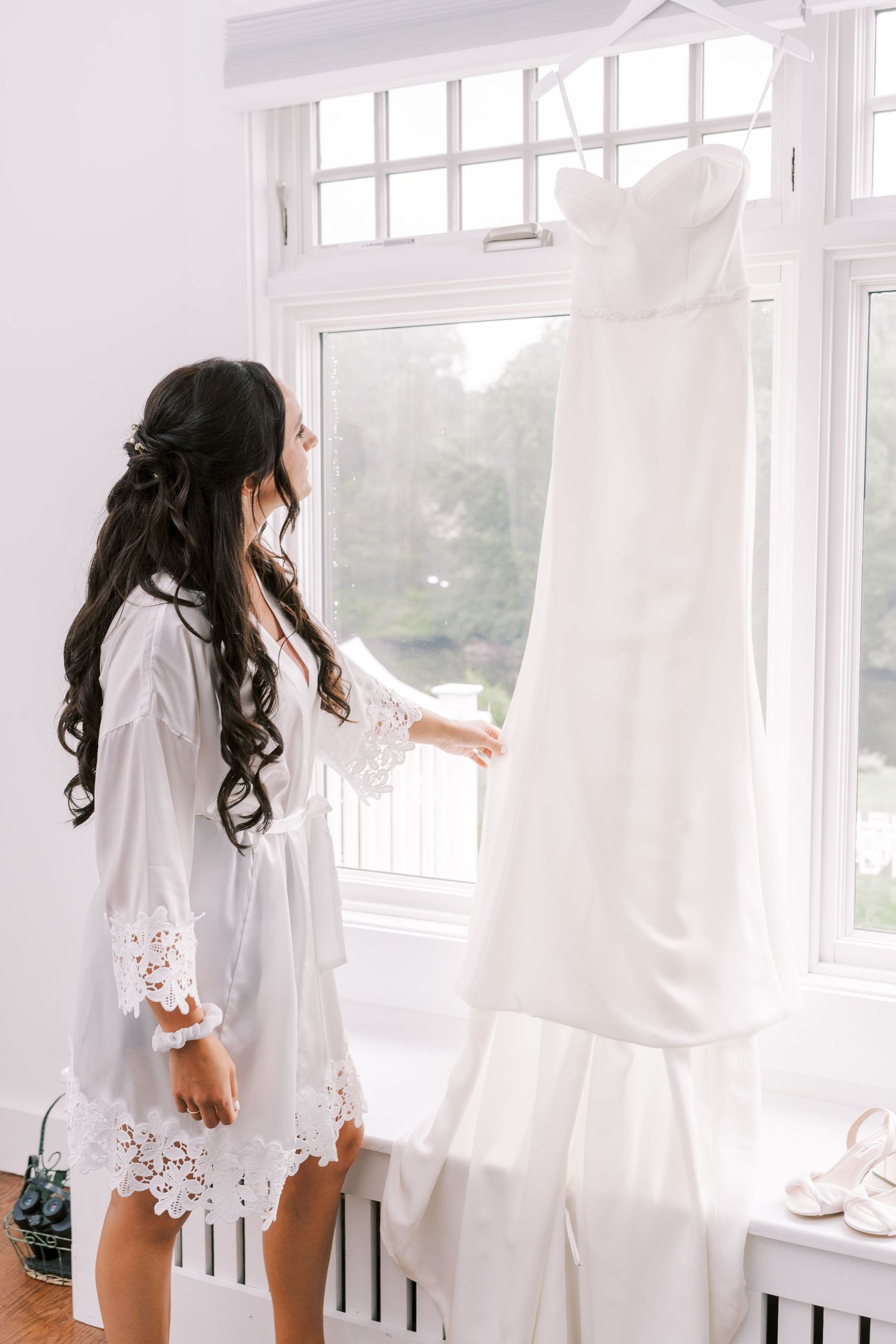 A white wedding dress hangs by a window while someone in a lace robe admires it during wedding preparations.