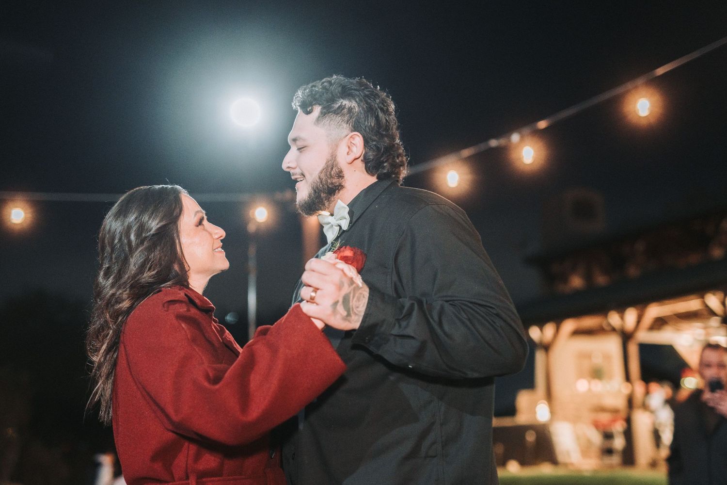 A couple dances together at night under string lights, with the person in red smiling at their dance partner.