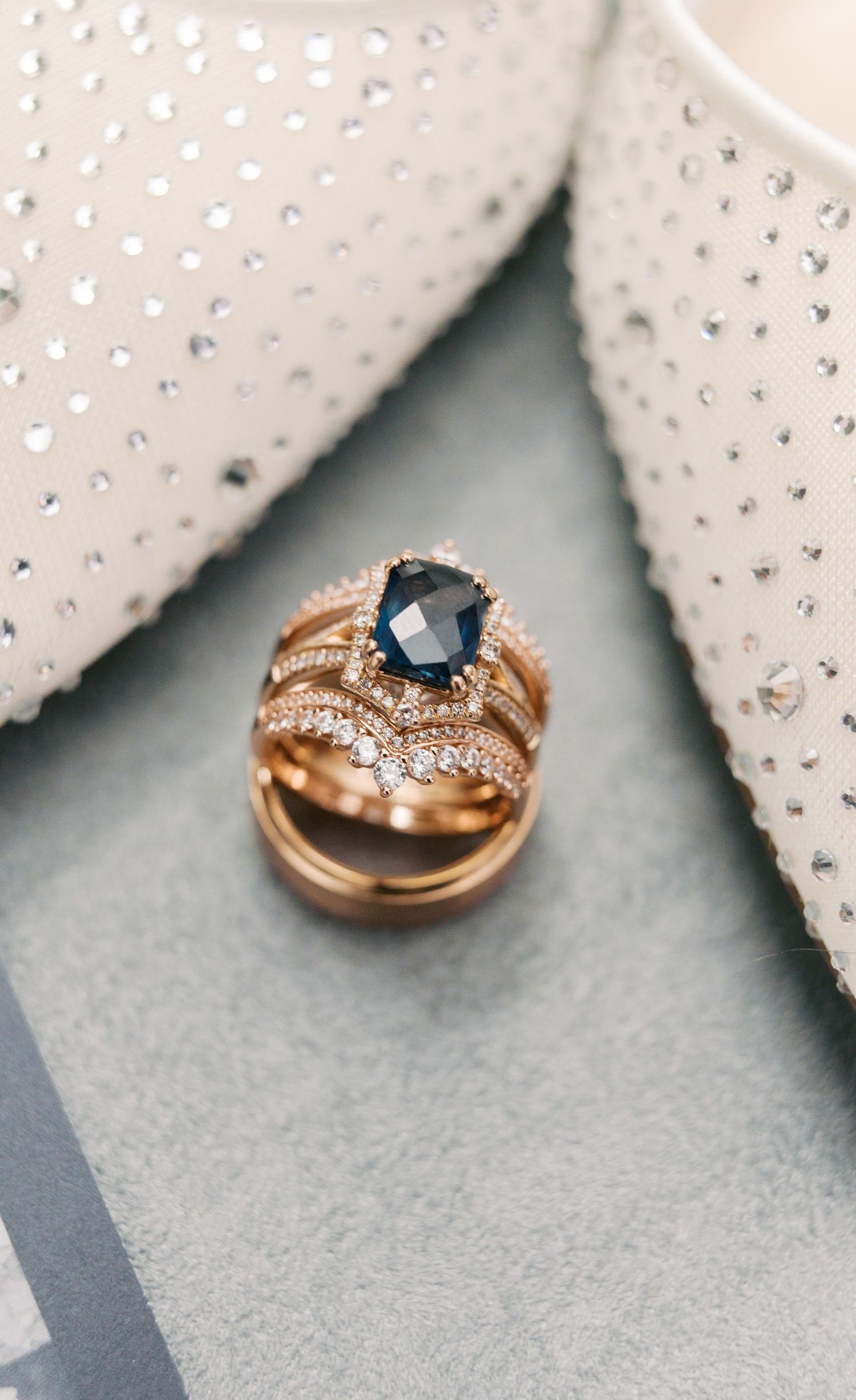 Rose gold engagement ring with dark blue sapphire center stone and diamond accents rests between sparkly wedding shoes.