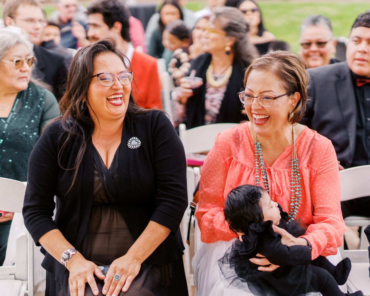 Two individuals share a joyful moment while sitting outdoors at an event, with one holding a small black dog.