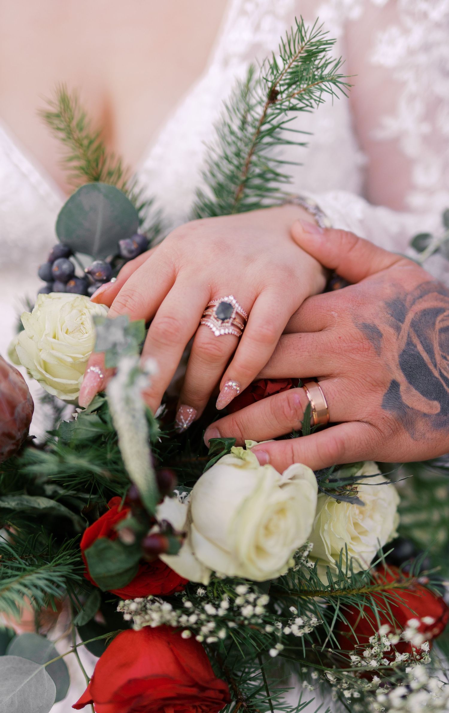 Two hands with rings and tattoos rest together on a winter wedding bouquet with red and white roses and evergreen sprigs.
