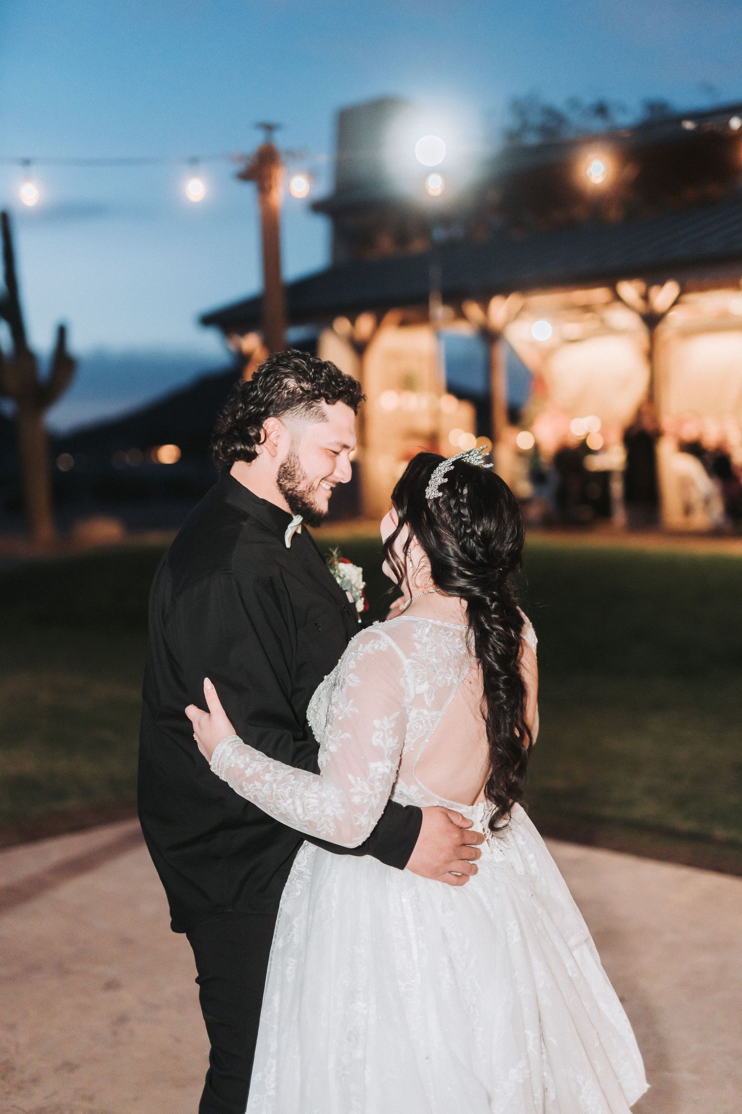 A couple shares an intimate moment while dancing outdoors at dusk under romantic string lights at their wedding reception.