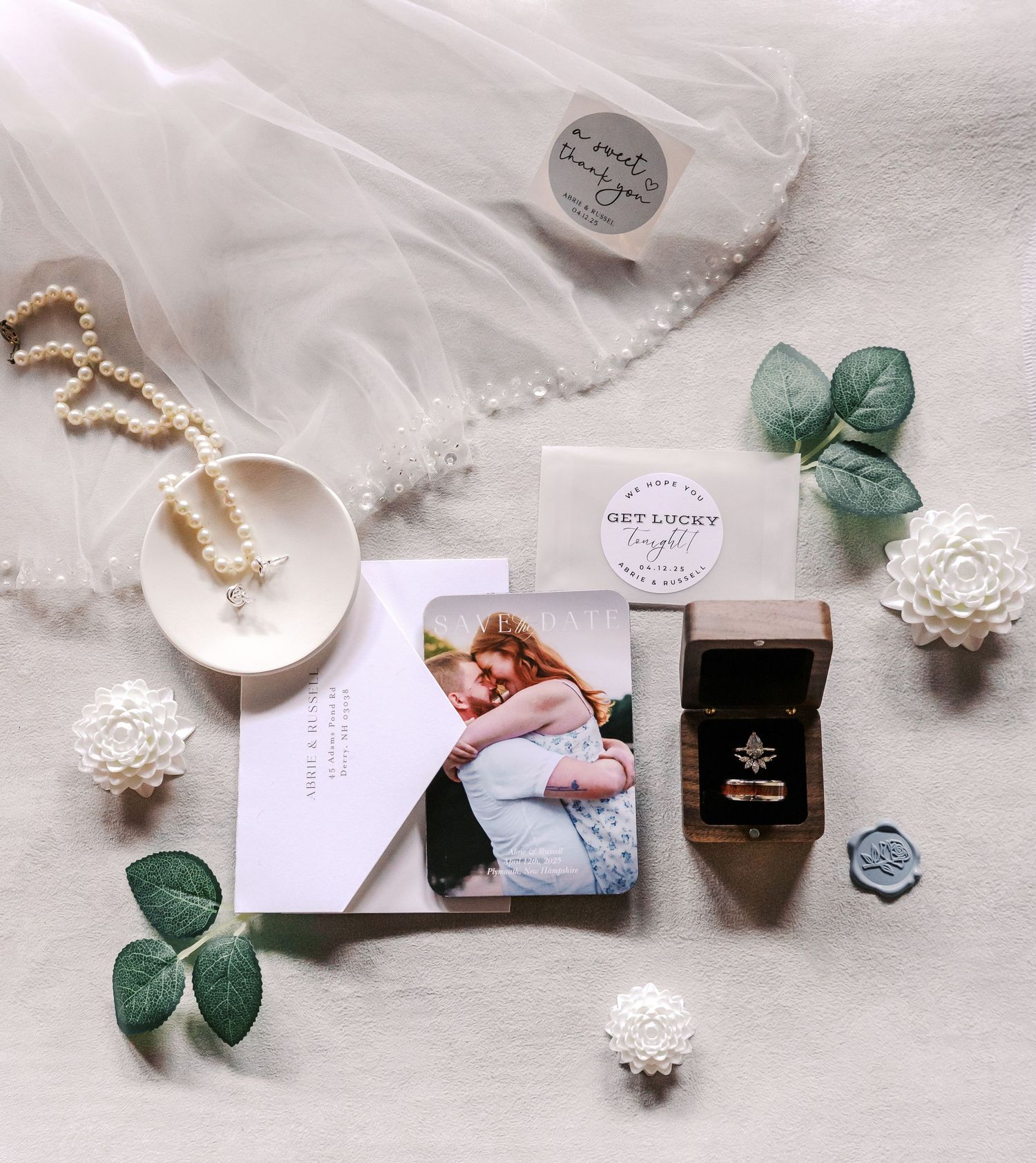 Wedding details flatlay with jewelry, greenery, and photo card on white fabric background.