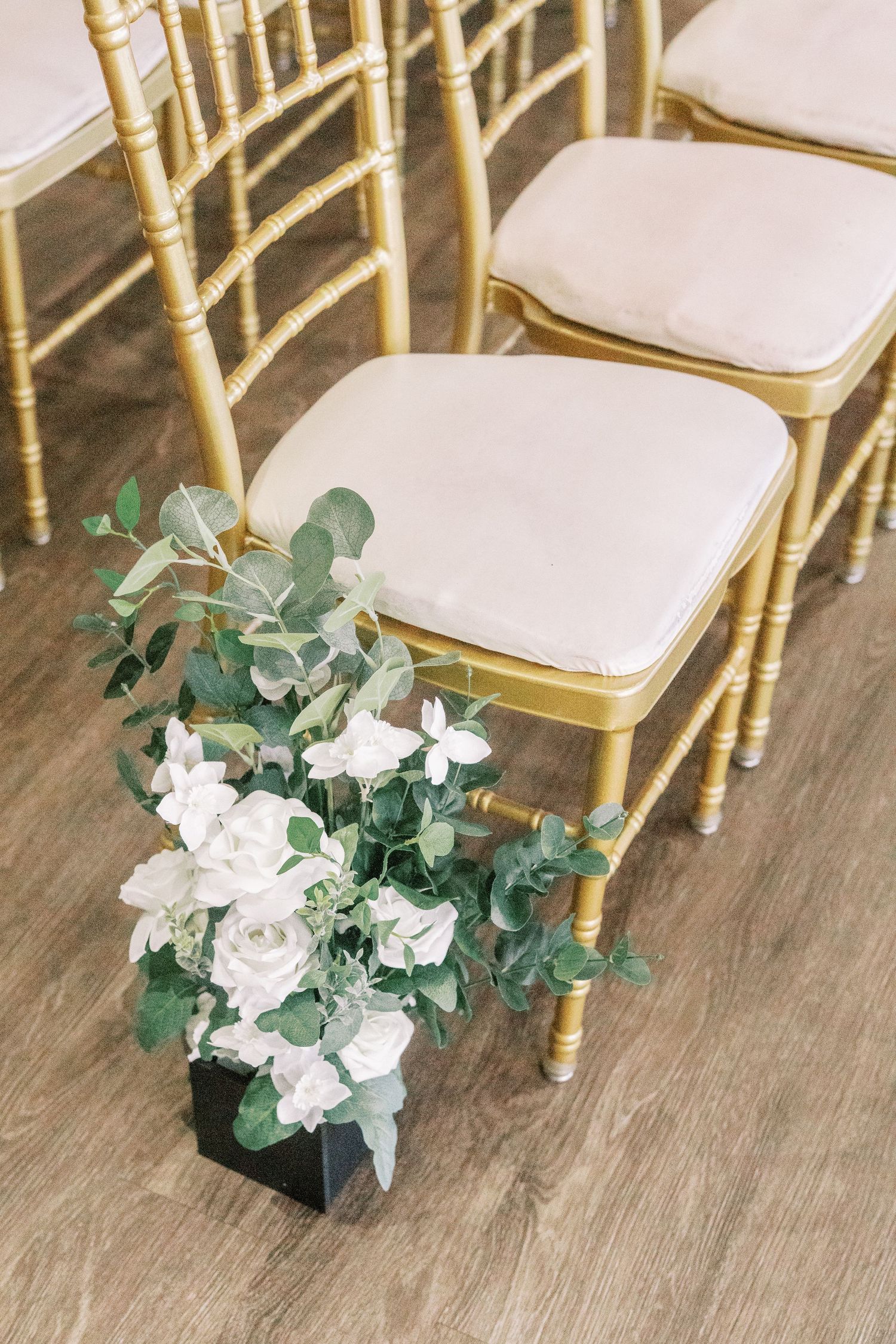Gold chiavari chairs with white cushions arranged with an elegant floral arrangement on hardwood floor.