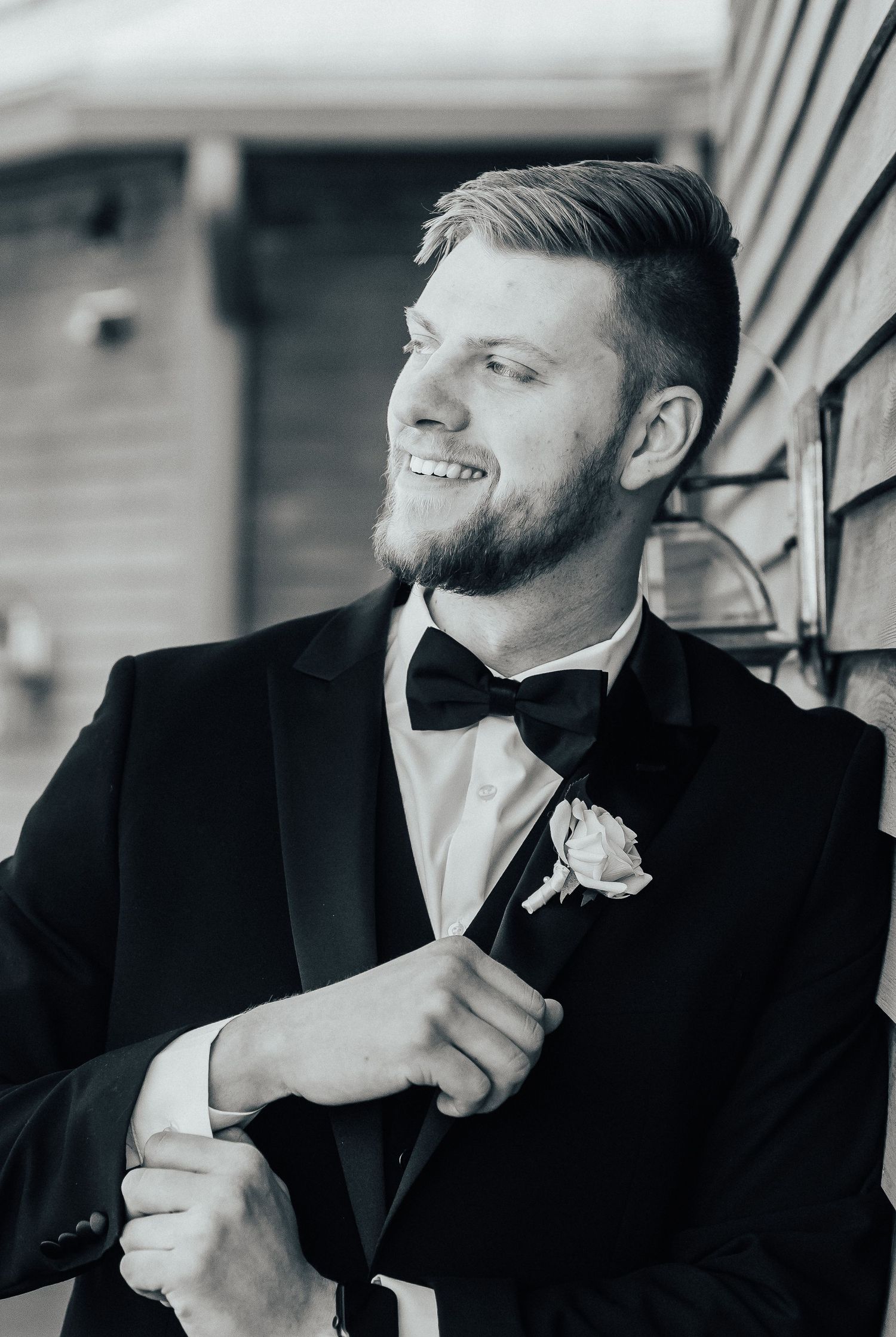 Black and white portrait of a groom in classic tuxedo and bow tie adjusting his boutonniere.