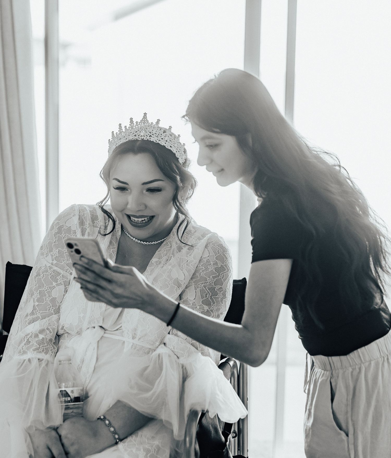 Black and white photo showing a bride in white dress and tiara looking at a phone screen while someone leans in beside her.