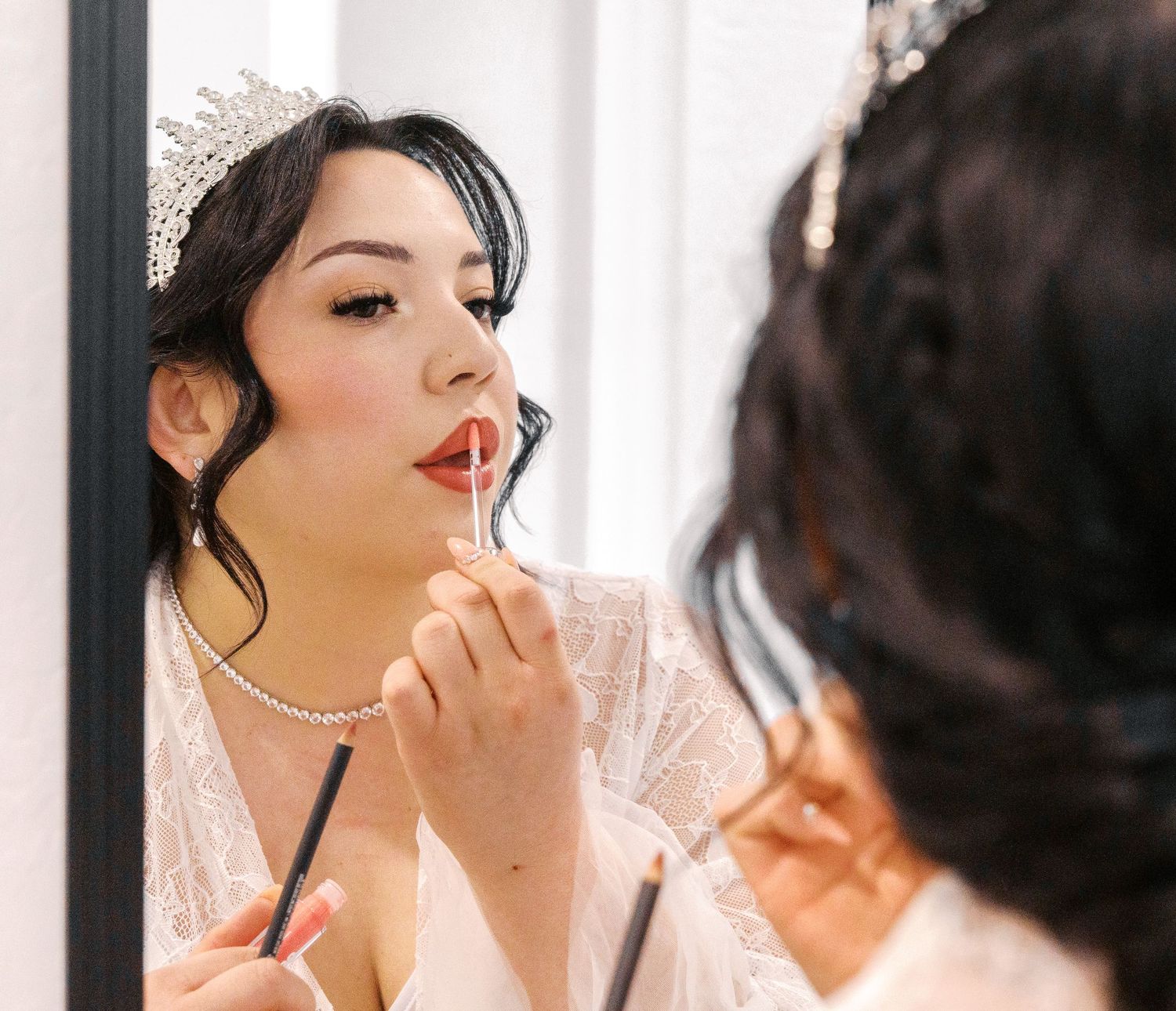Someone applies lipstick while looking in a mirror, wearing a pearl necklace and light-colored robe.